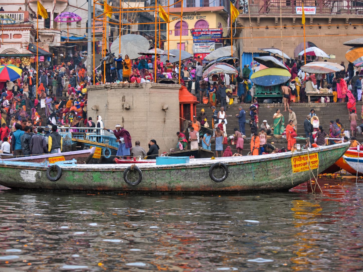 A long weathered green boat fills the foreground beneath crowded ghat steps, umbrellas and riverfront structures in Varanasi, 2019