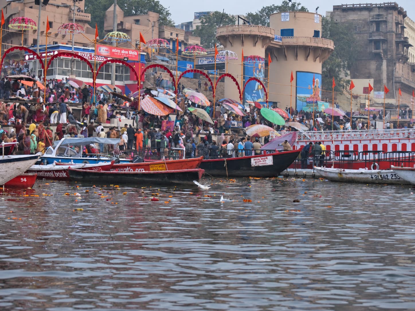 A broad river view shows crowded ghat steps, patterned umbrellas, moored boats and layered riverfront buildings in Varanasi, 2019