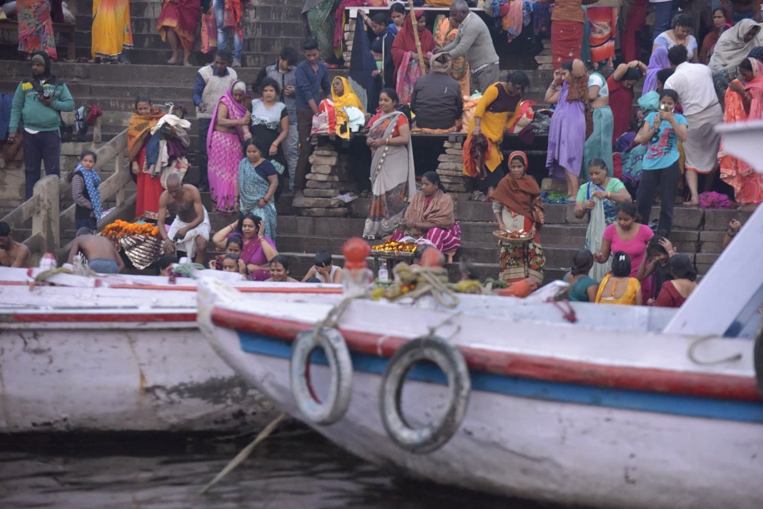 Working boats fill the foreground while a dense crowd gathers on the ghat steps behind them in Varanasi, 2019