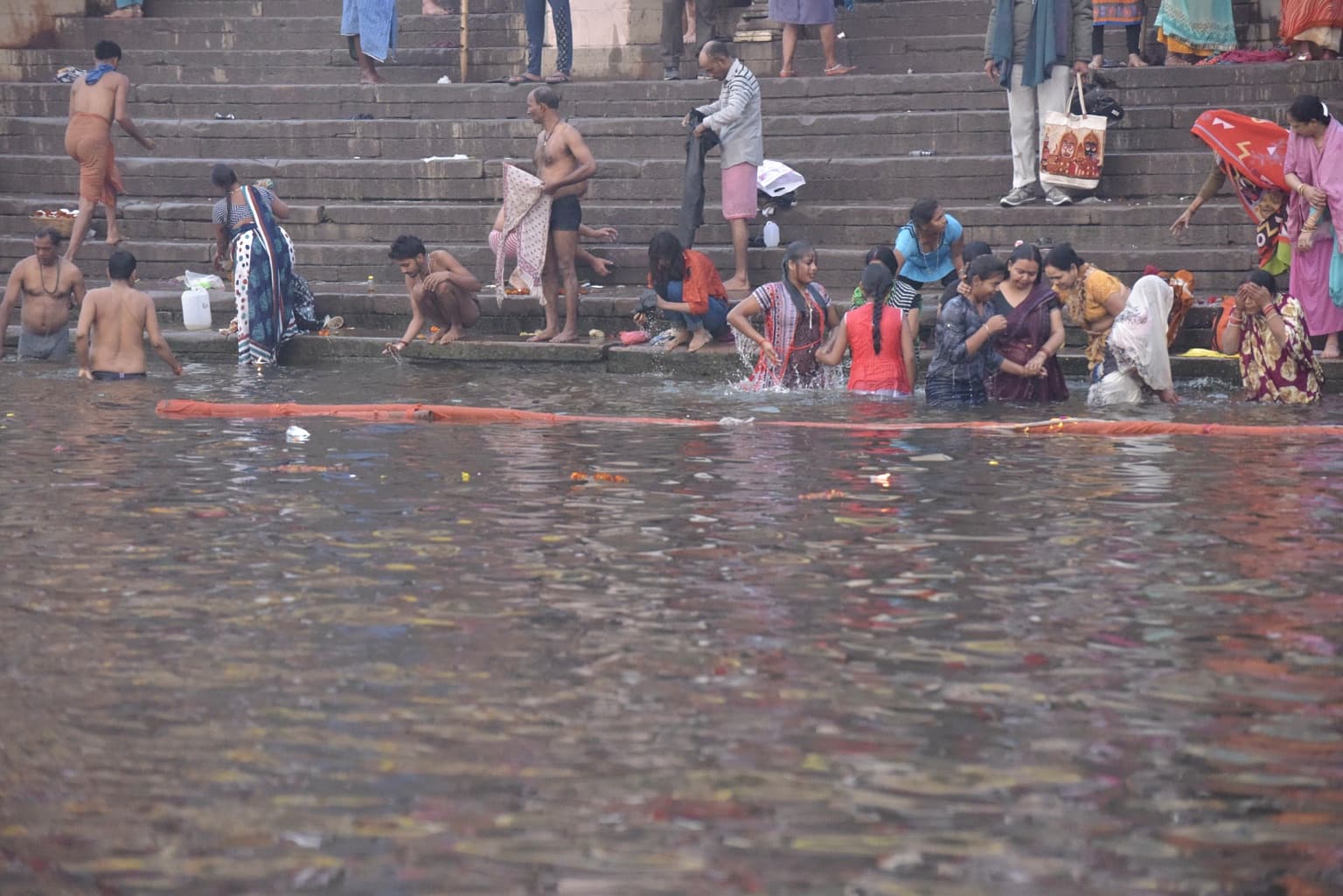 A group of bathers enters and splashes in the Ganges while others stand and dress on the stone steps behind them in Varanasi, 2019