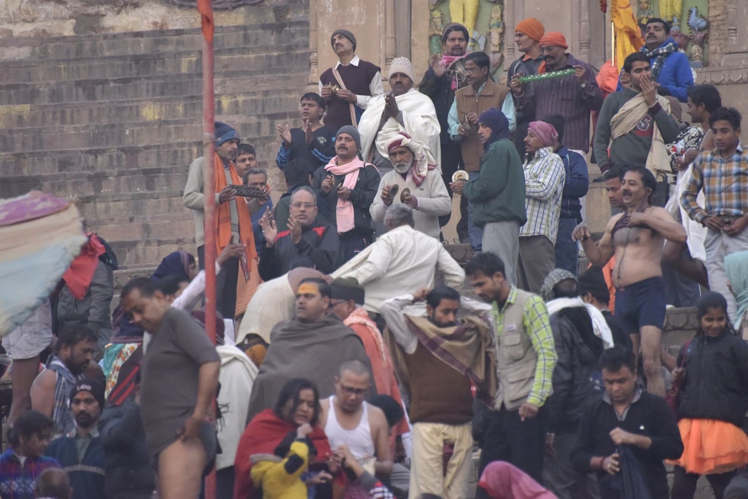 A group of men gathers on the ghats with hand cymbals and other devotional objects amid the wider riverfront crowd in Varanasi, 2019