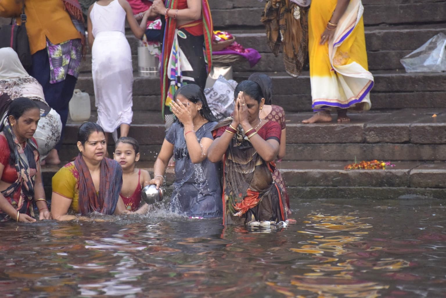 Women stand waist-deep at the Ganges as one presses her hands together and another splashes water over her face, Varanasi, 2019