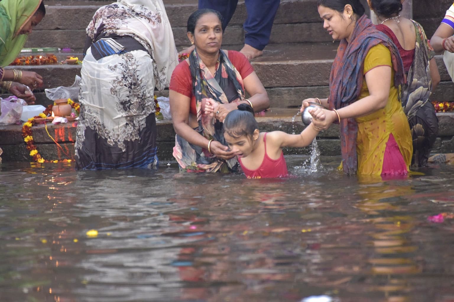 A young girl is supported by two women as she lowers herself into the Ganges at the foot of the ghats in Varanasi, 2019