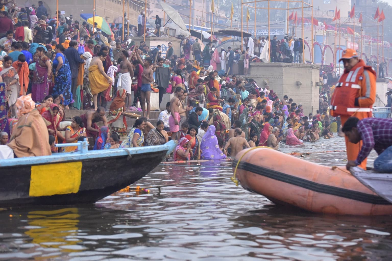 A dense crowd bathes, washes and gathers along the riverfront while boats frame the foreground in Varanasi, 2019