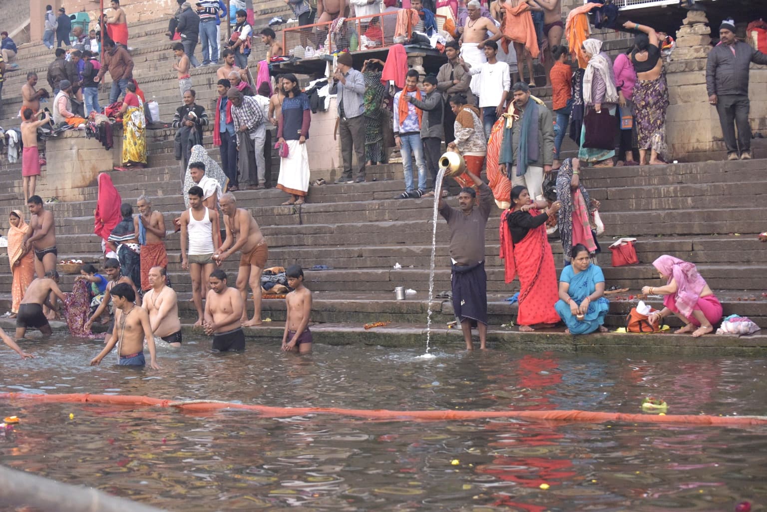 A man pours water from a brass vessel while bathers and families gather along the lower ghat steps in Varanasi, 2019