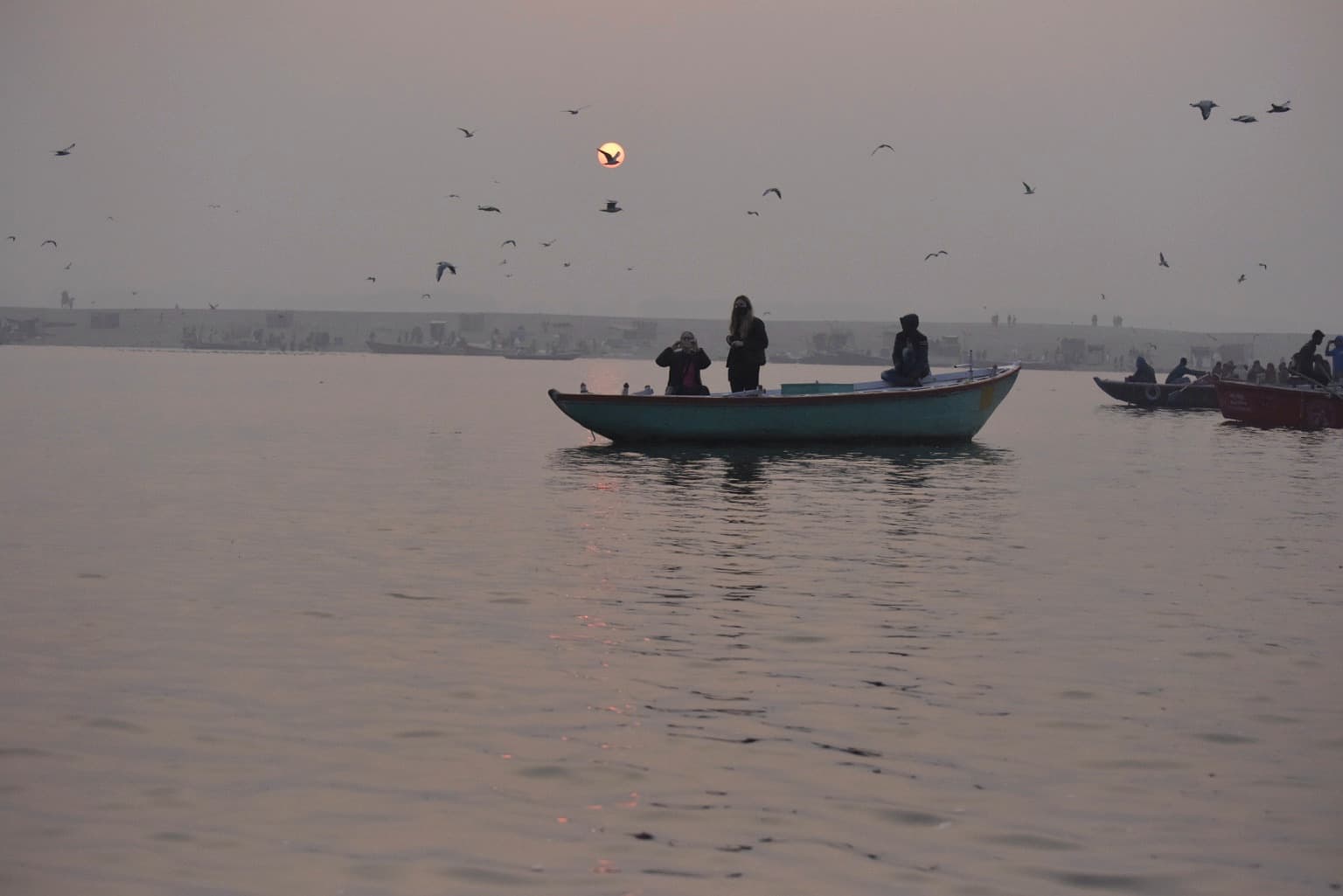 A passenger boat crosses the Ganges in soft sunrise light while birds move through the hazy sky above Varanasi, 2019