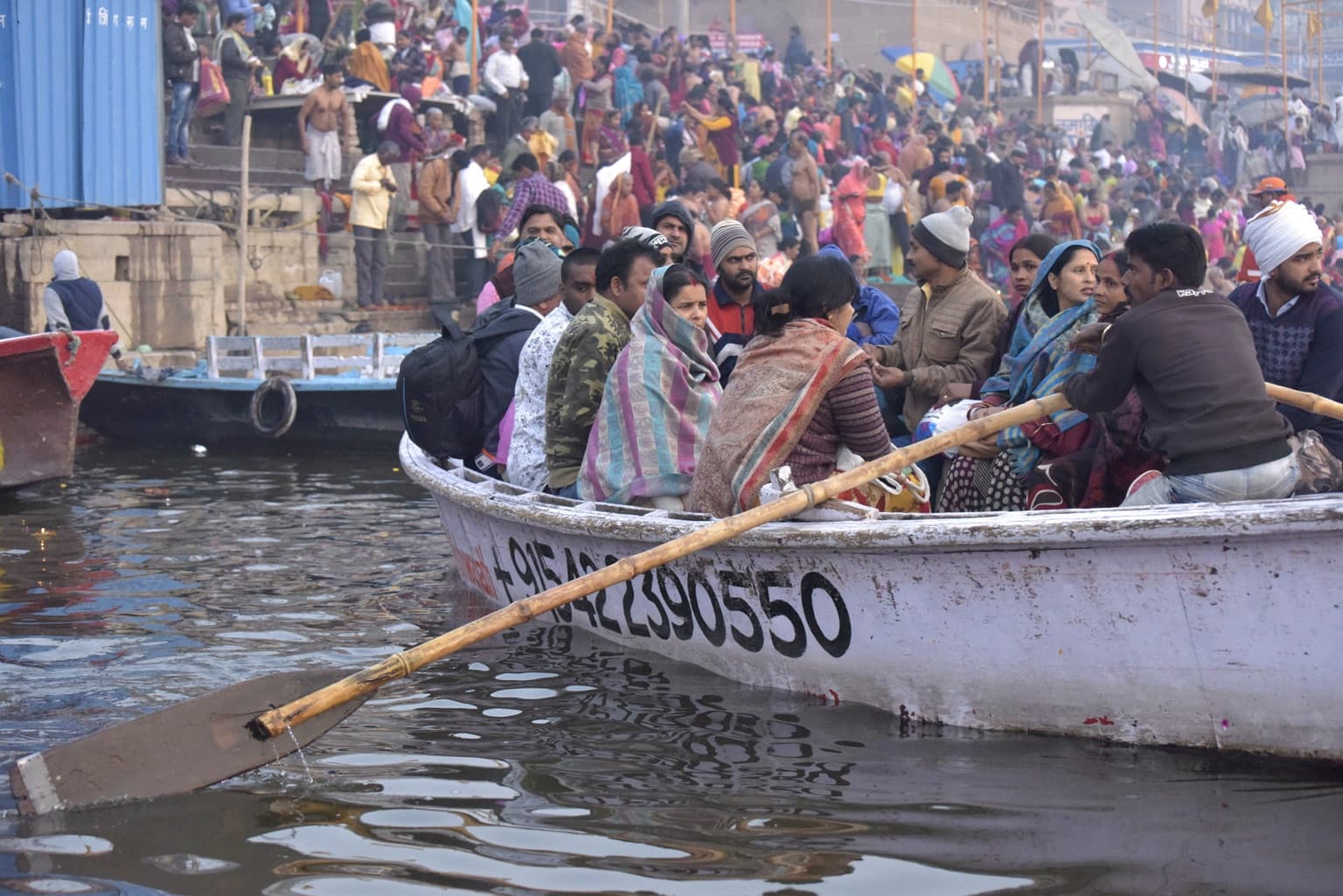 Passengers crowd a low wooden boat close to the camera while the busy ghats rise behind them in Varanasi, 2019