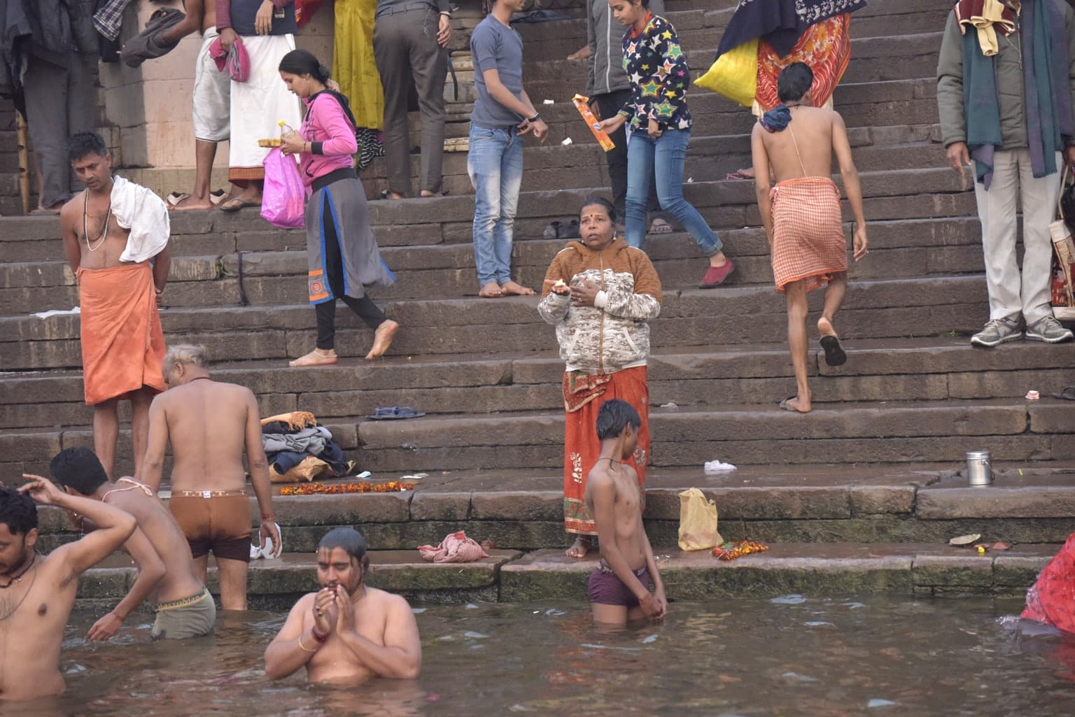 Bathers and worshippers occupy the lower steps beside the Ganges as a woman stands holding a small offering in Varanasi, 2019