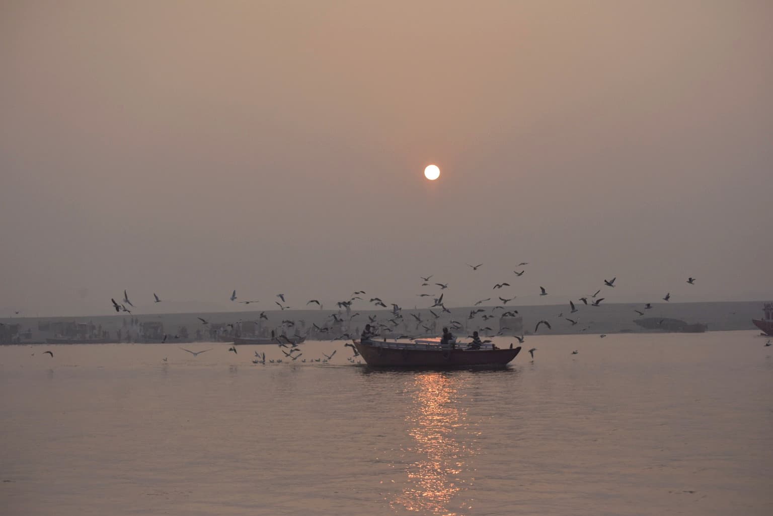 A small boat passes beneath a pale rising sun while birds circle above the Ganges in the morning haze of Varanasi, 2019