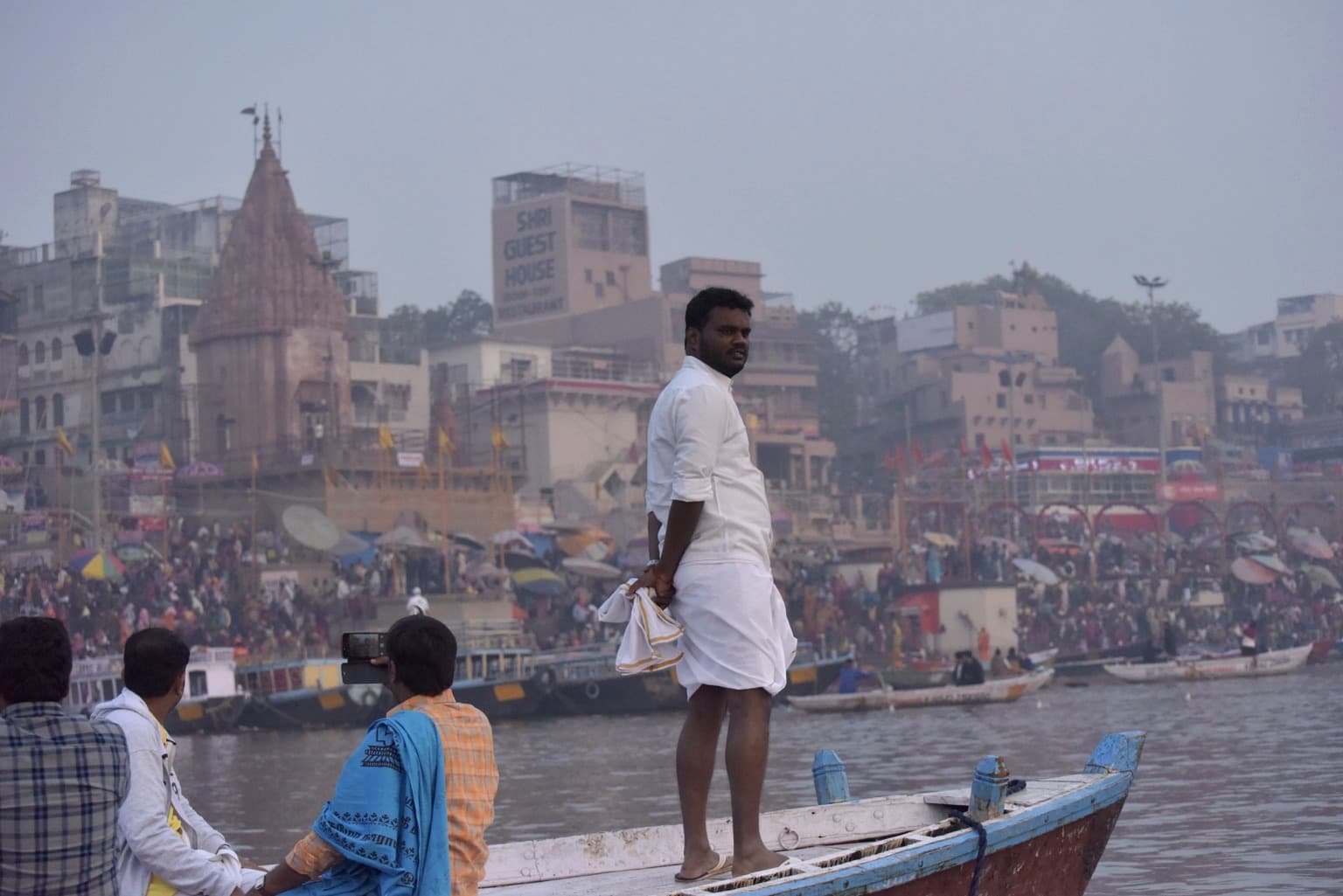 A man in white stands upright in a small boat on the Ganges while the crowded ghats rise behind him in the morning haze of Varanasi, 2019