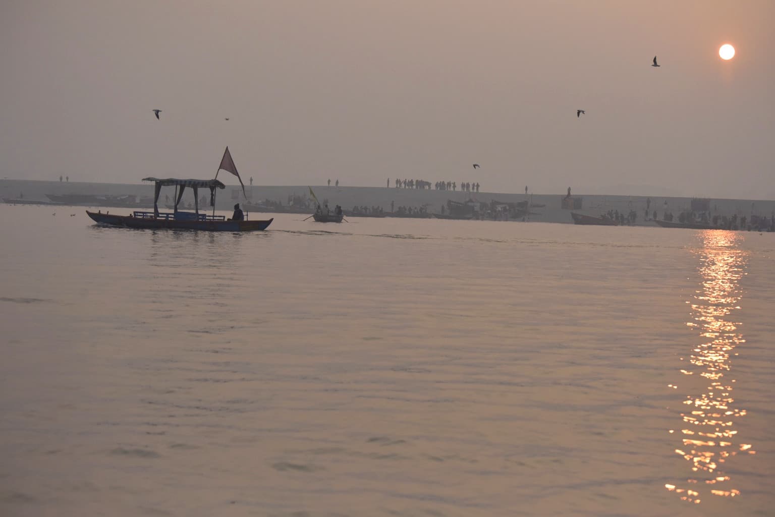 A low sun glows over the Ganges while small boats and birds cross the warm morning haze in Varanasi, 2019