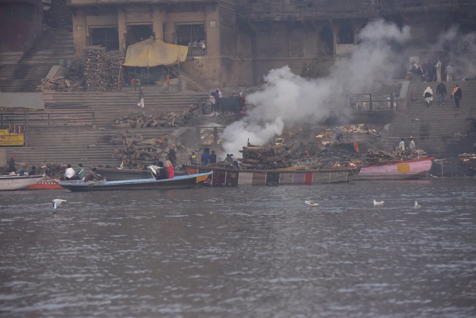 Smoke rises above stacked cremation wood and working boats along the riverfront cremation ghat in Varanasi, 2019