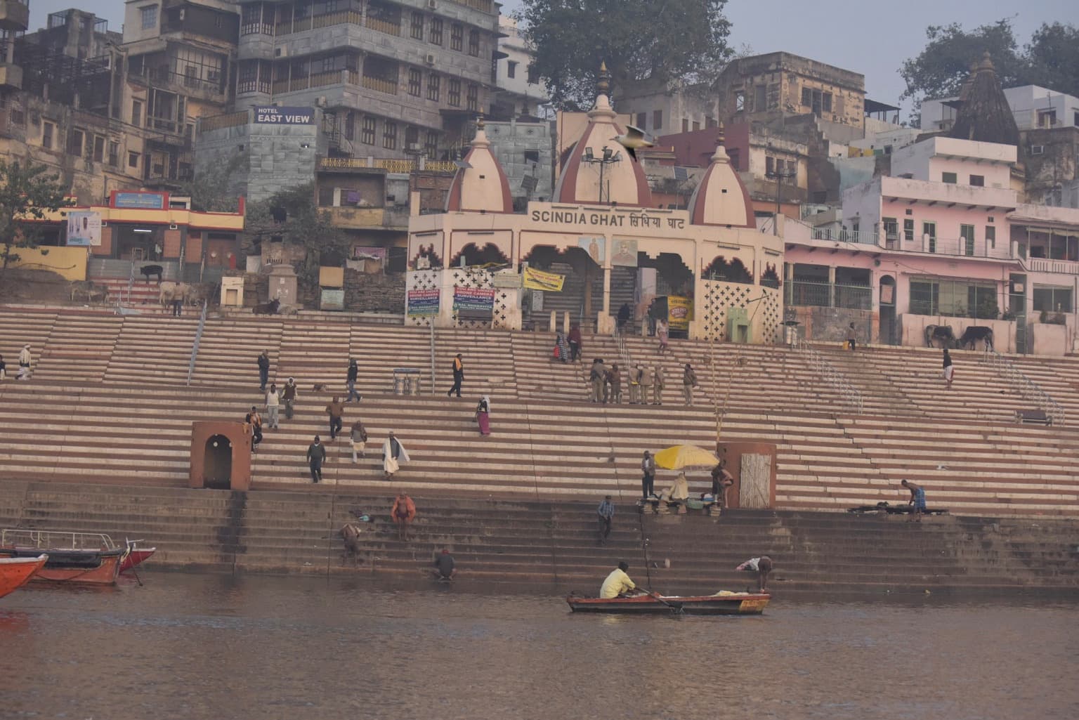 A broad view of striped ghat steps, a riverfront shrine and scattered figures beside the Ganges in Varanasi, 2019