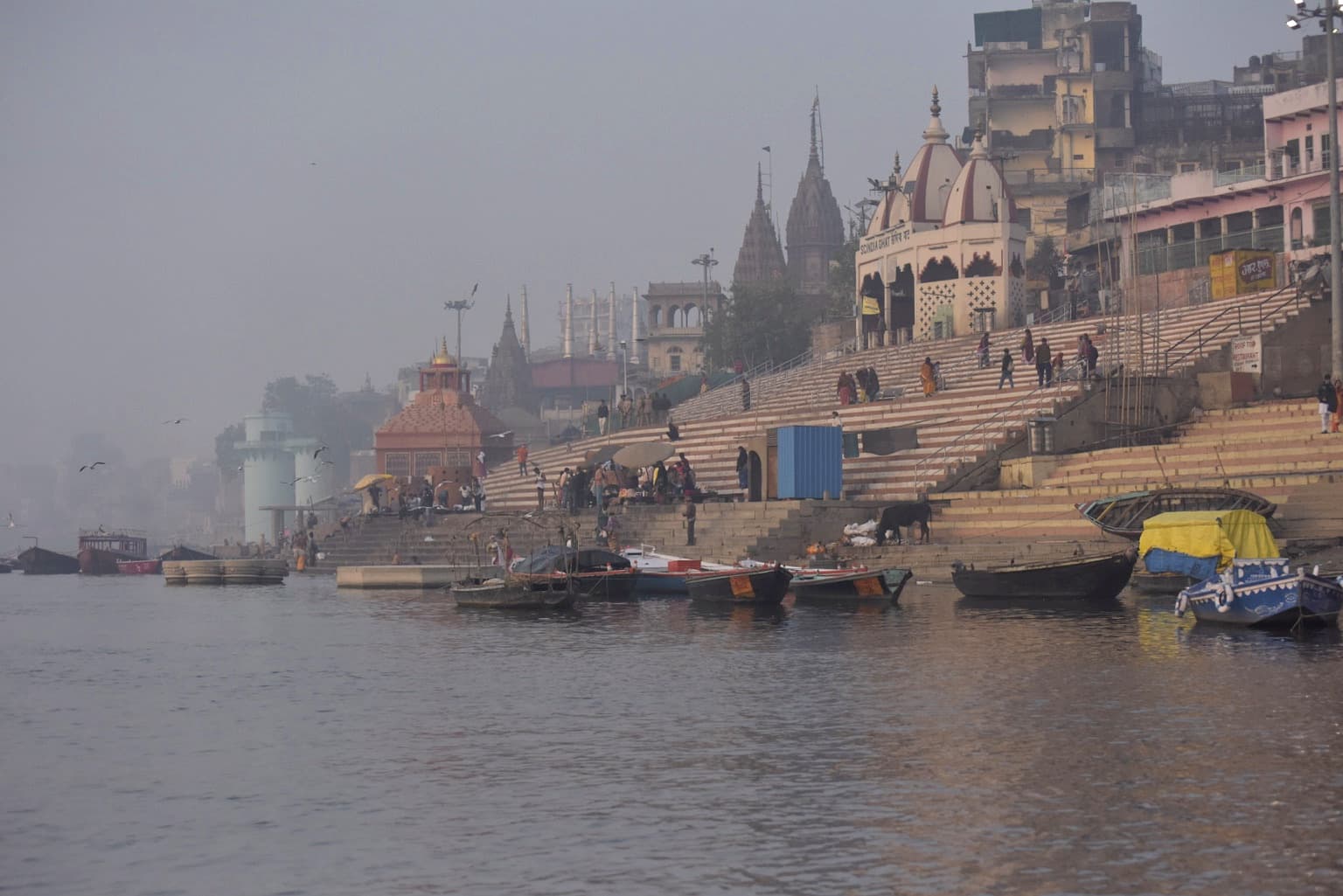 A long oblique view follows stepped ghats, moored boats and temple forms into the morning haze beside the Ganges in Varanasi, 2019