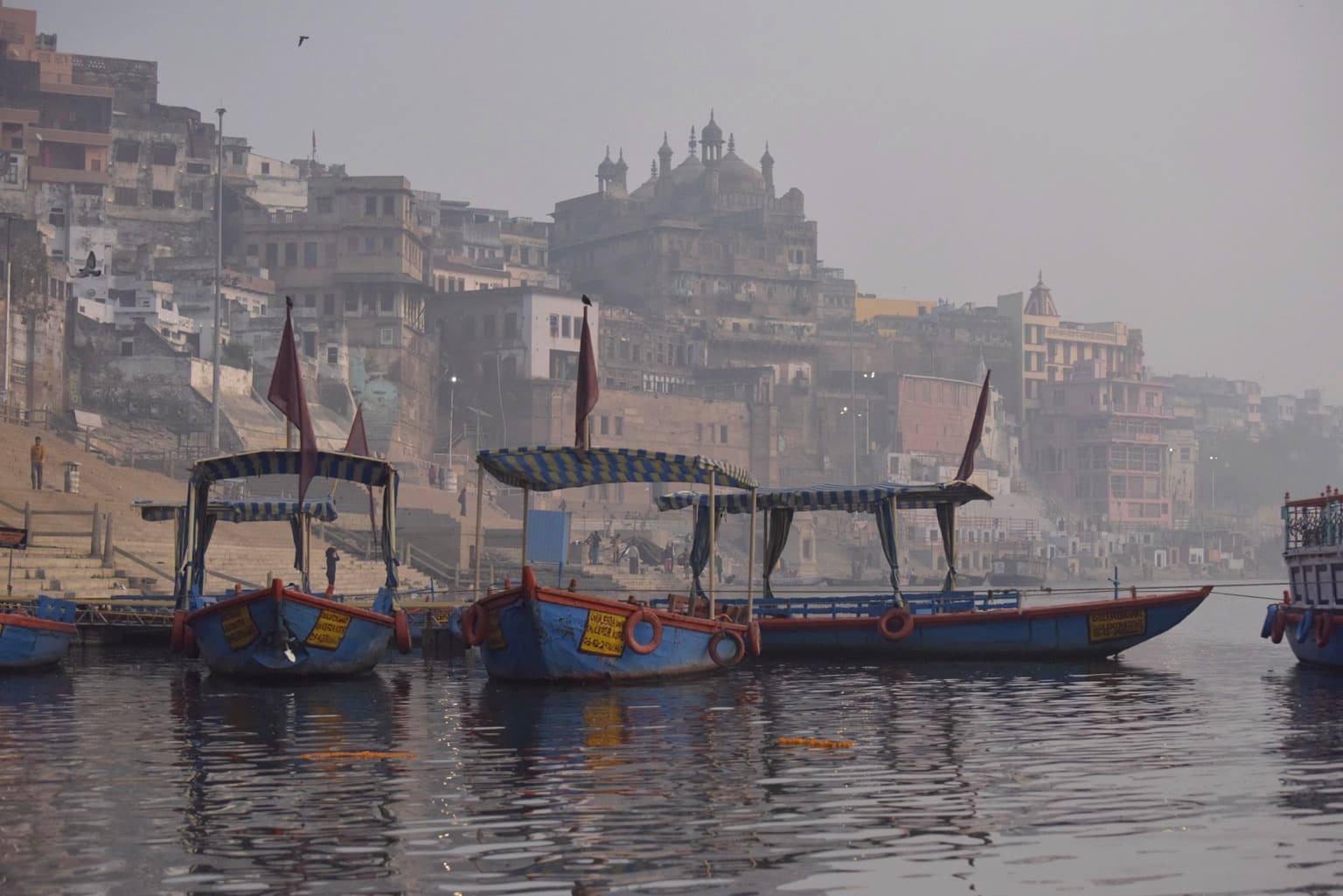 Moored wooden boats sit before layered riverfront buildings in the morning haze of Varanasi, 2019