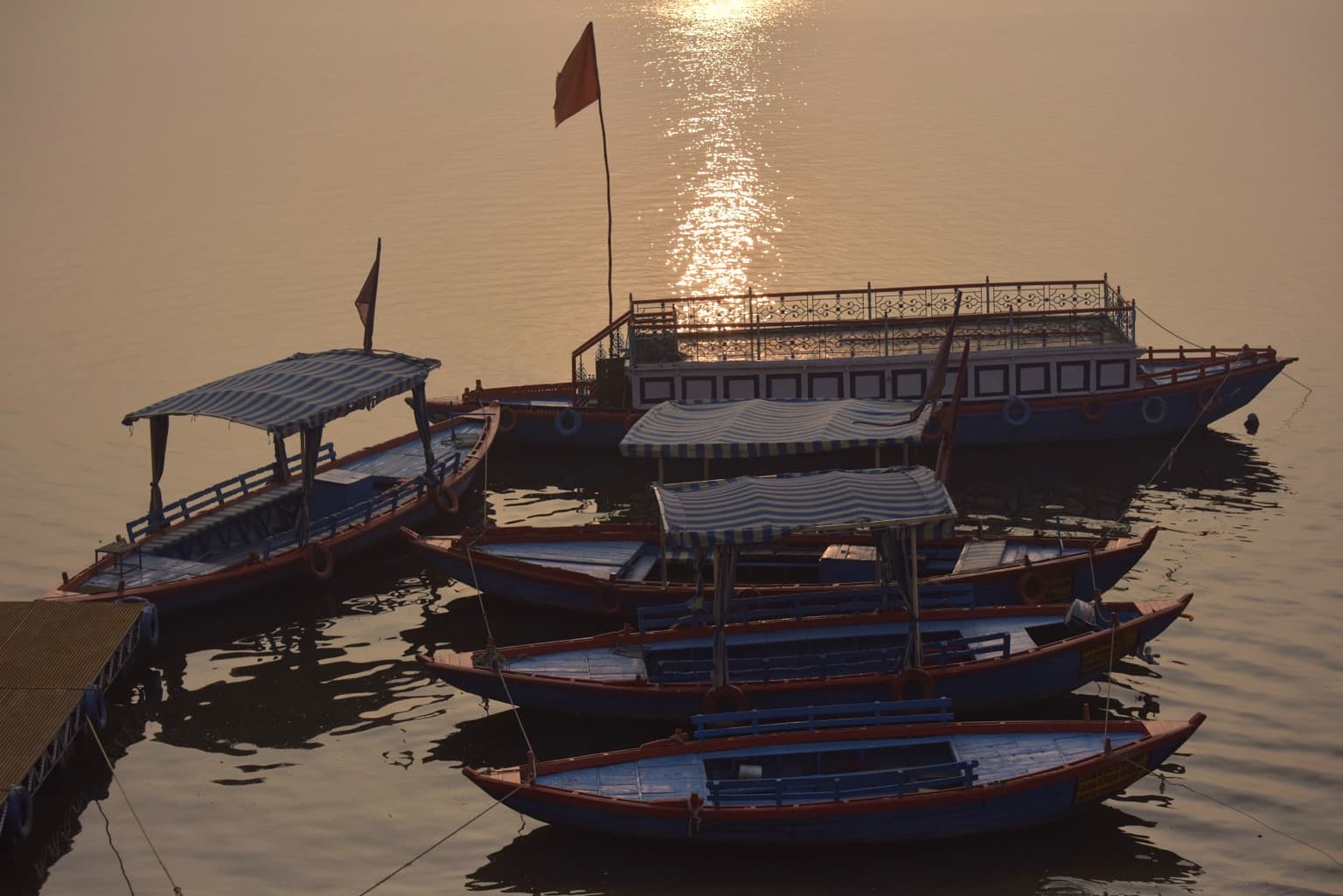 Several blue and red passenger boats cluster beneath a warm column of reflected sunlight on the Ganges in Varanasi, 2019