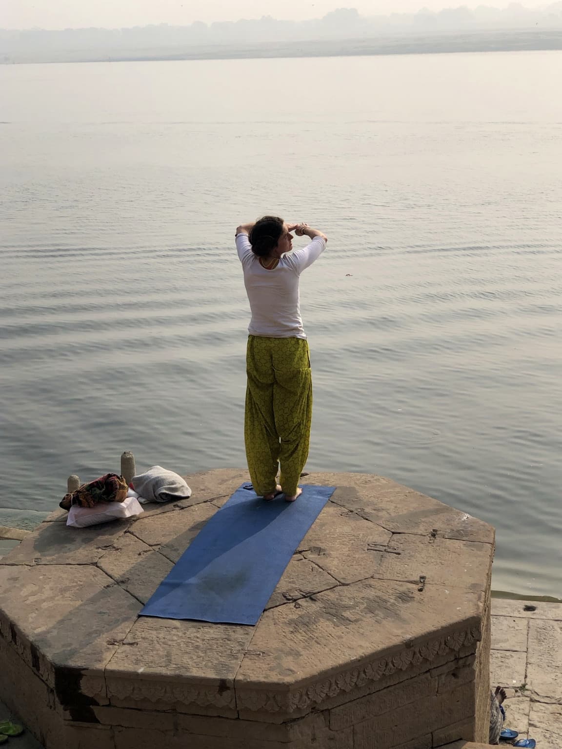 A woman in a white top and green trousers stands on a blue exercise mat on a stone platform above the Ganges in Varanasi, 2019