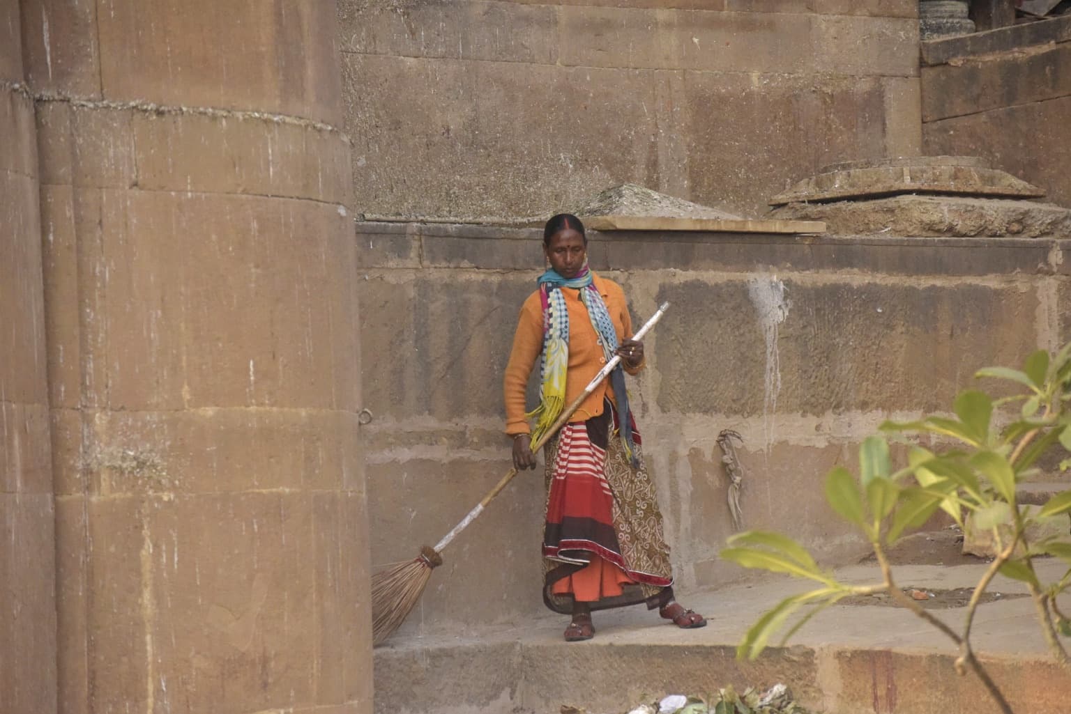 A woman in orange clothing sweeps beside monumental stone walls on the ghats of Varanasi, 2019