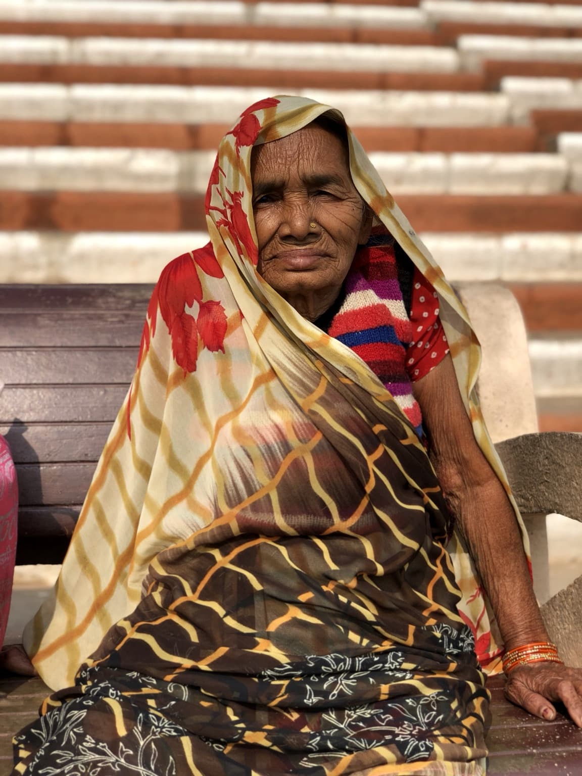 An elderly woman in a patterned sari sits for a direct portrait on the broad striped steps of the ghats in Varanasi, 2019