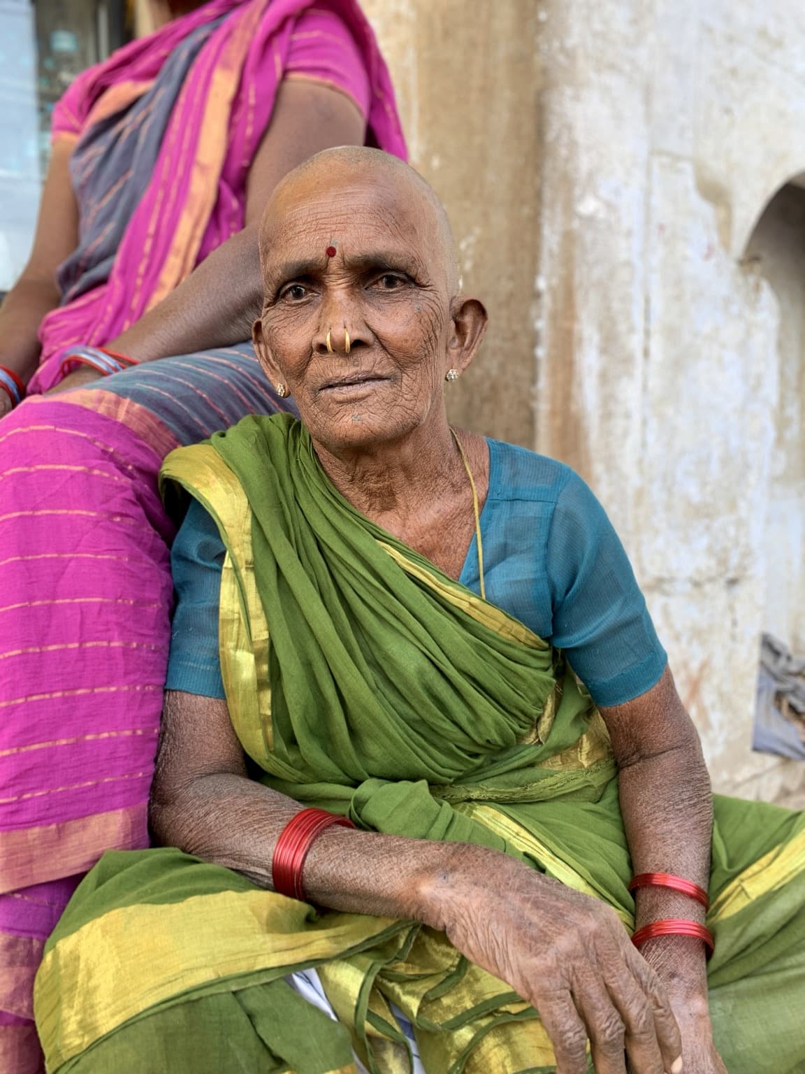 An elderly woman in a green sari and blue blouse looks directly toward the camera in Varanasi, 2019