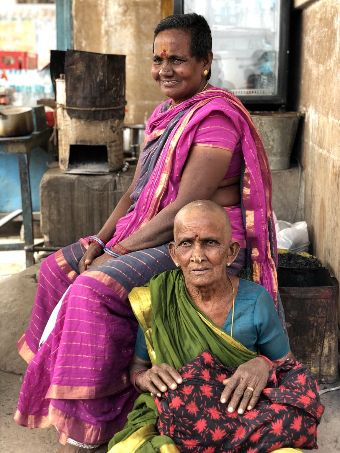 Two women in green and magenta clothing sit together beside a weathered riverfront wall in Varanasi, 2019