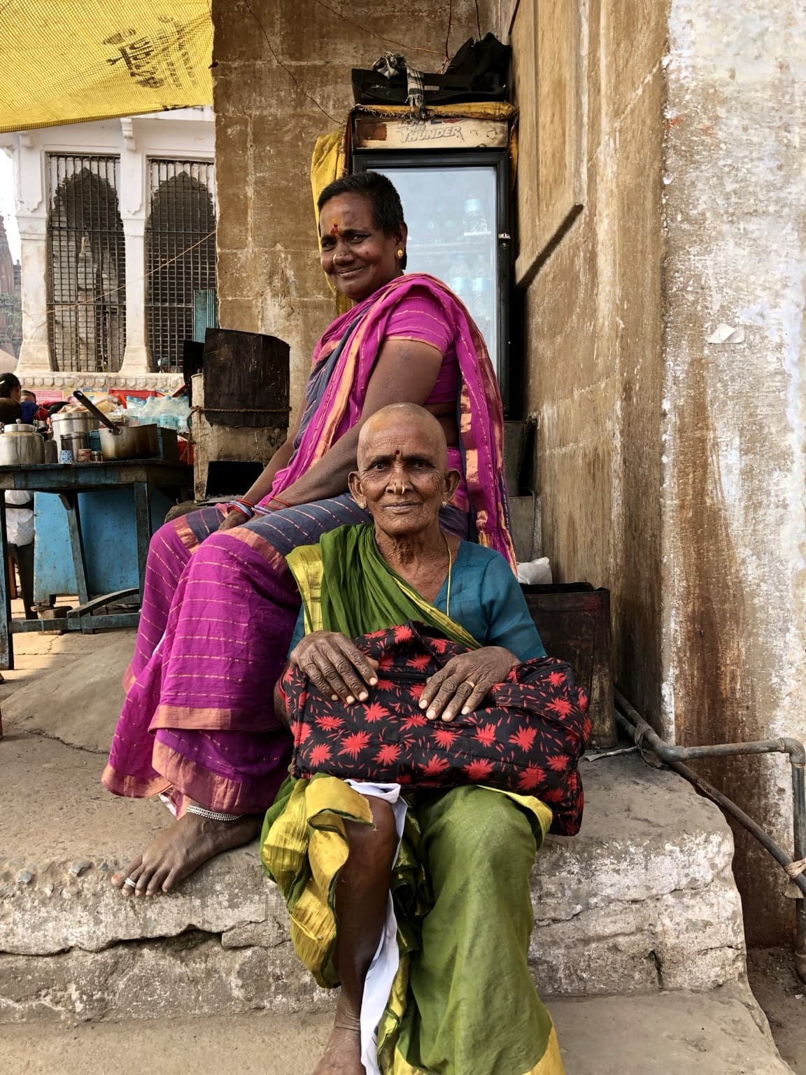 Two women in green and magenta saris sit together in a narrow riverfront setting in Varanasi, 2019