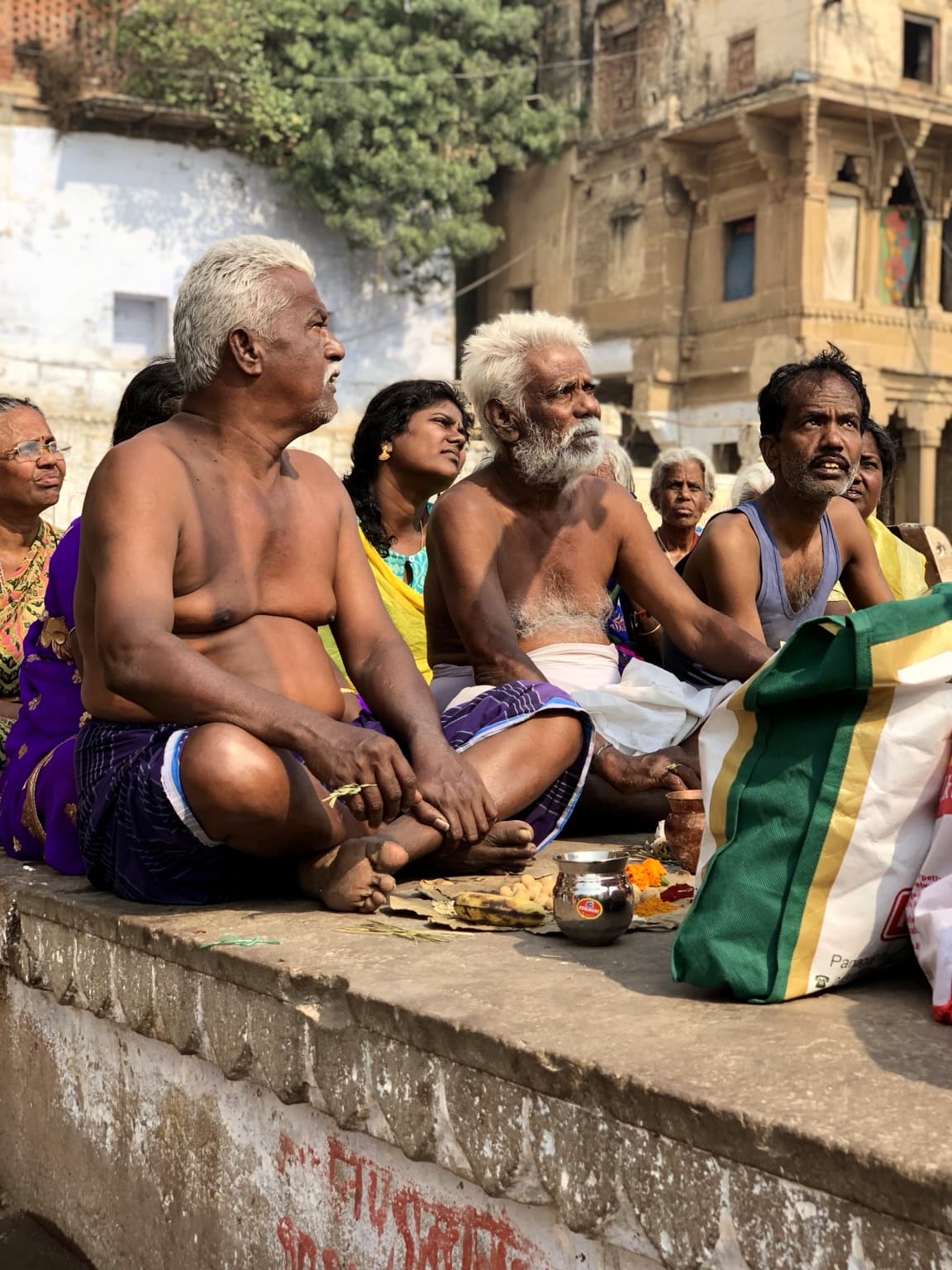 Men and women sit closely together around small vessels and offerings during a riverfront gathering in Varanasi, 2019
