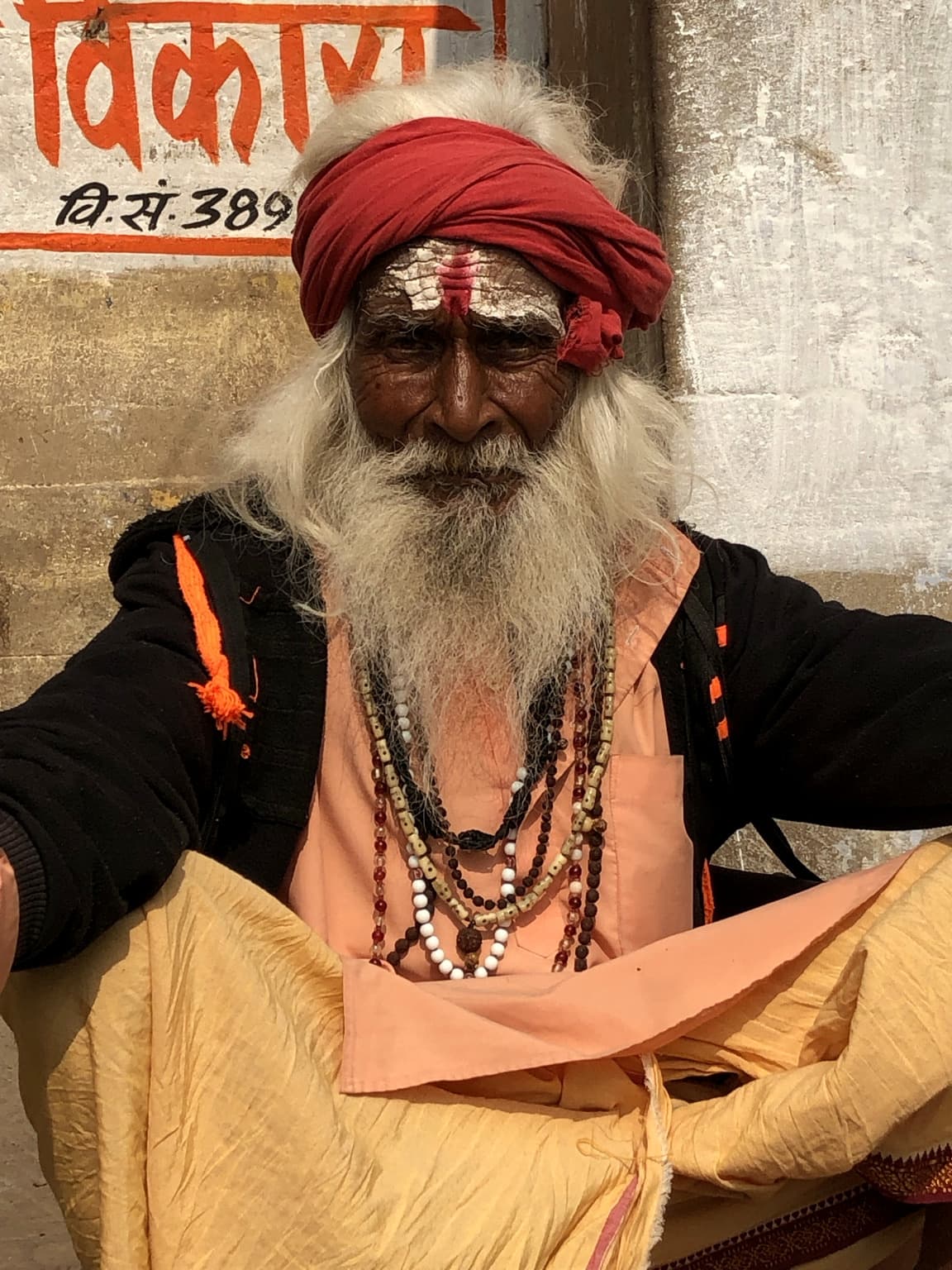 An elderly sadhu with a red turban, long white beard and painted forehead sits facing the camera in Varanasi, 2019