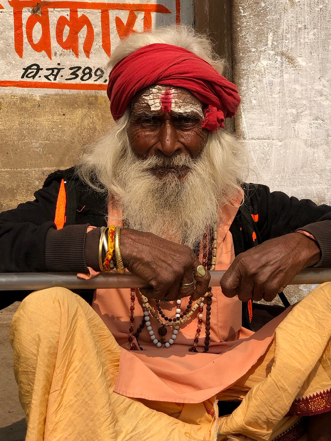 An elderly sadhu in a red turban rests both hands on a staff while looking directly toward the camera in Varanasi, 2019