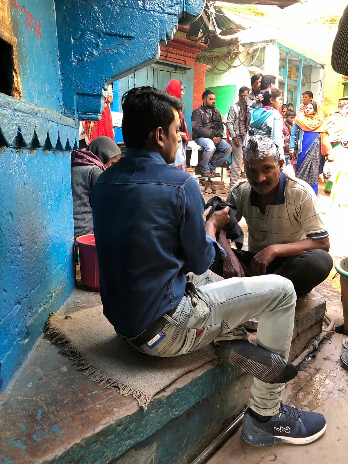 A barber washes a seated man’s hair in a narrow blue-painted working lane while people gather behind them in Varanasi, 2019