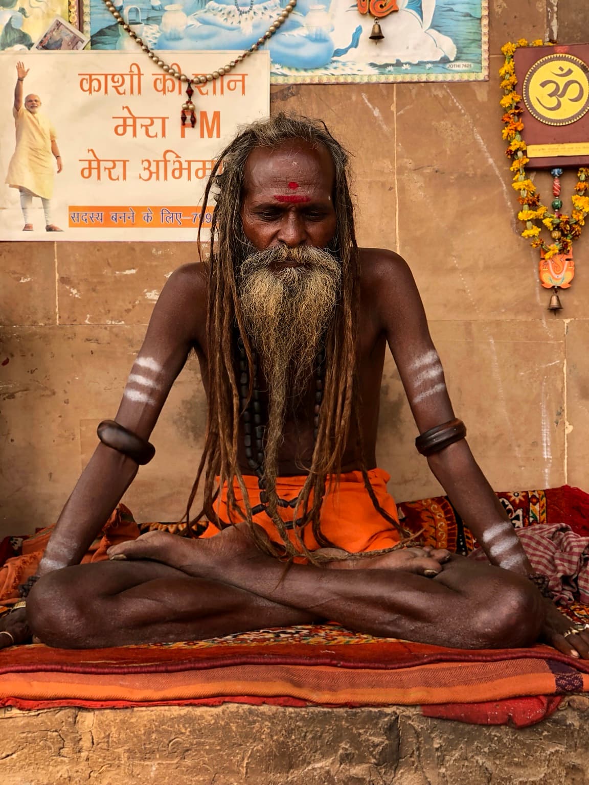 A long-haired sadhu sits cross-legged in meditation with white markings on his arms and orange cloth at his waist in Varanasi, 2019