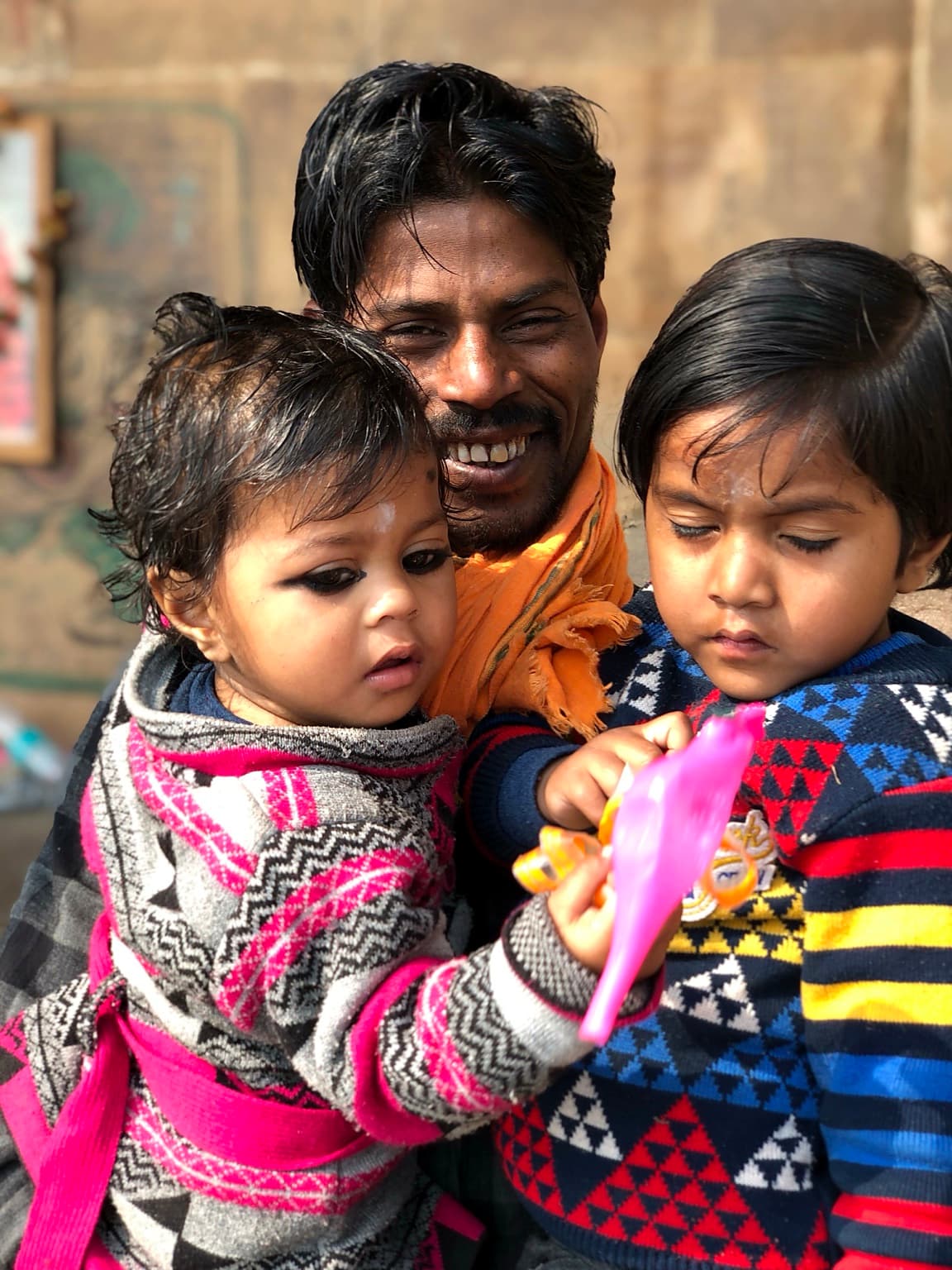 Two young children focus on a bright toy while their smiling father holds them close in Varanasi, 2019