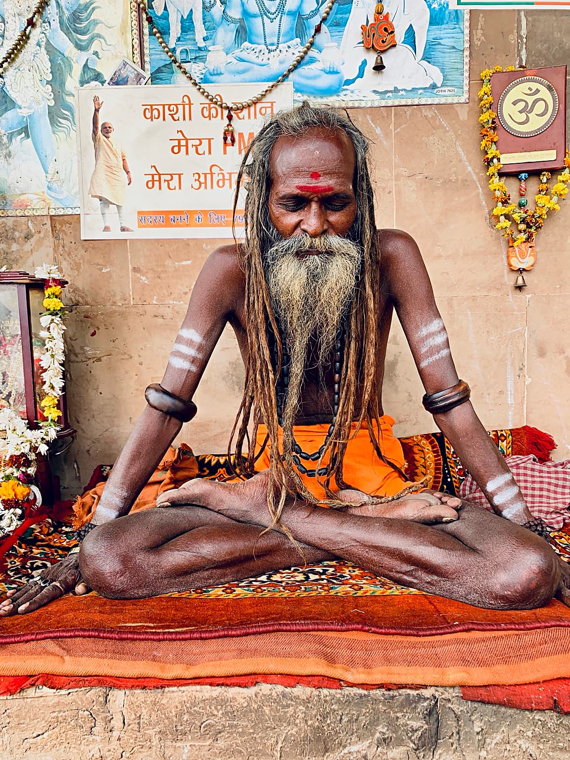 A bearded sadhu with ash markings and closed eyes sits amid religious imagery on the ghats of Varanasi, 2019