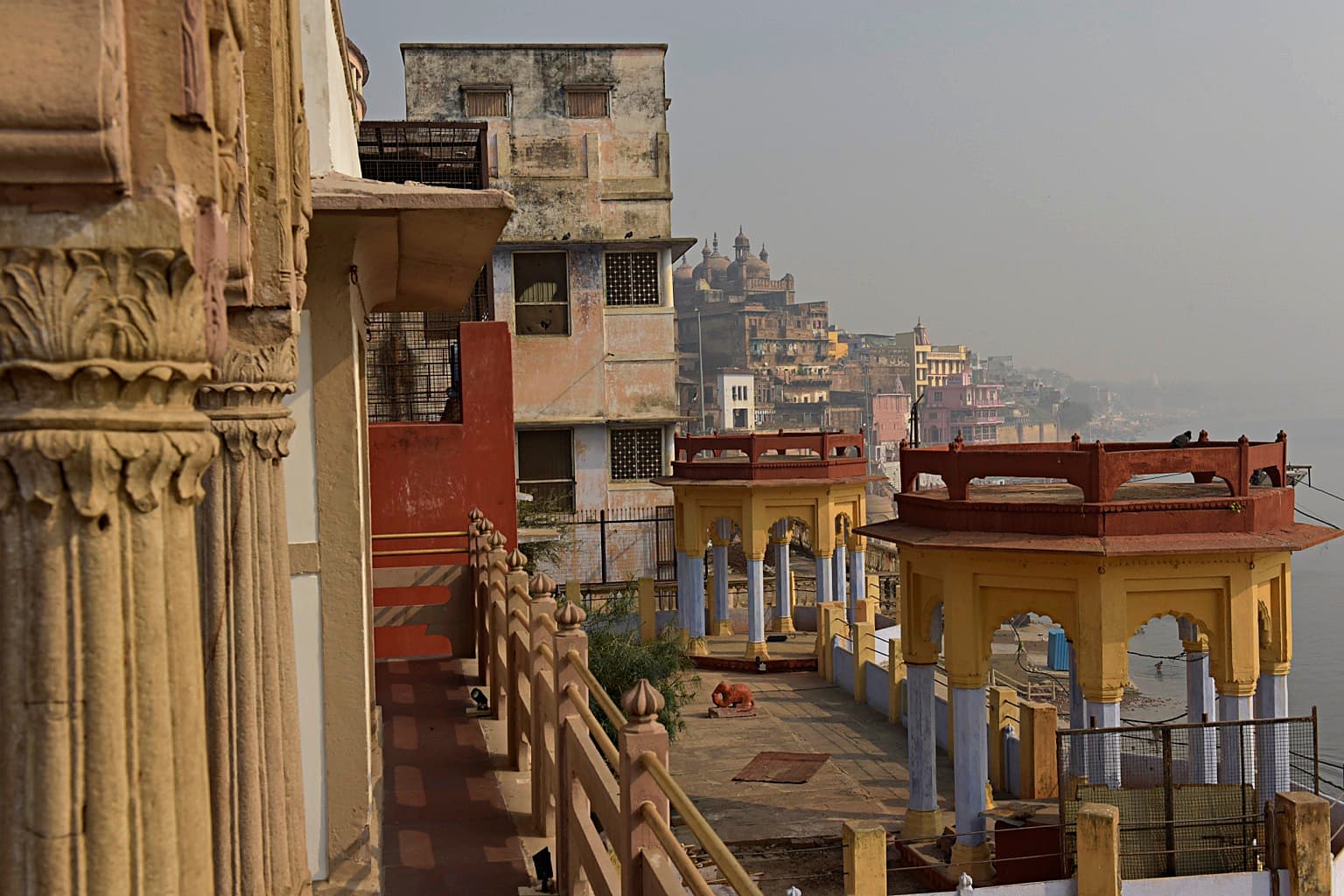 A carved stone column frames repeating riverfront pavilions and the hazy Ganges in Varanasi, 2019