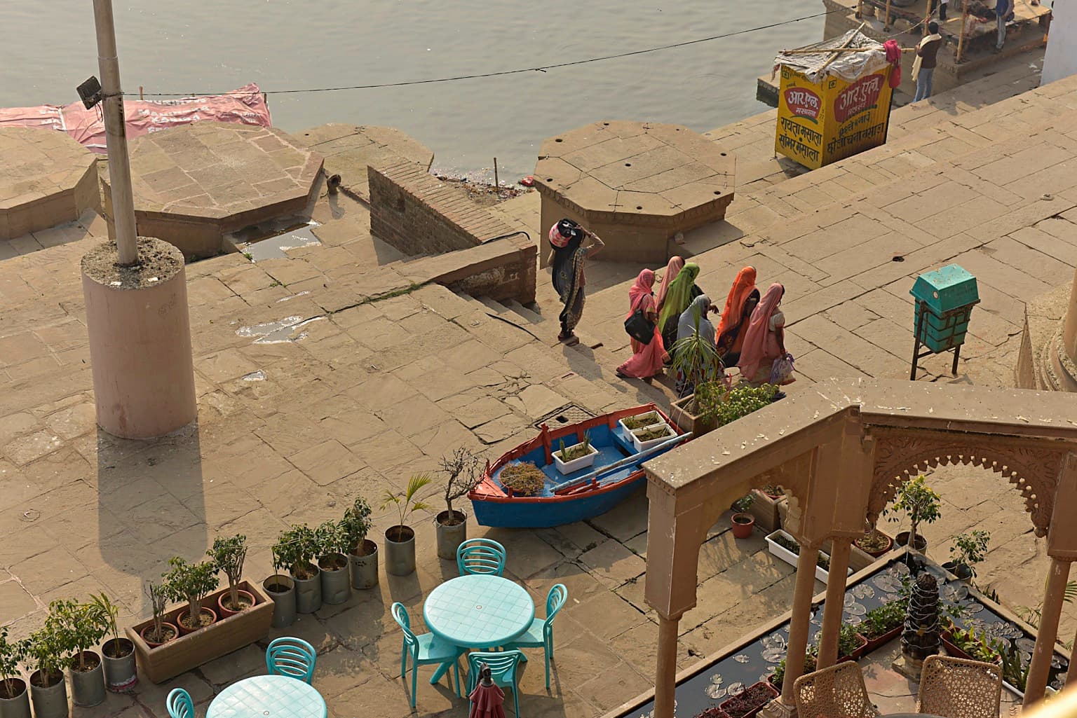Women and other figures move through an elevated view of daily life on the ghats beside a blue boat in Varanasi, 2019