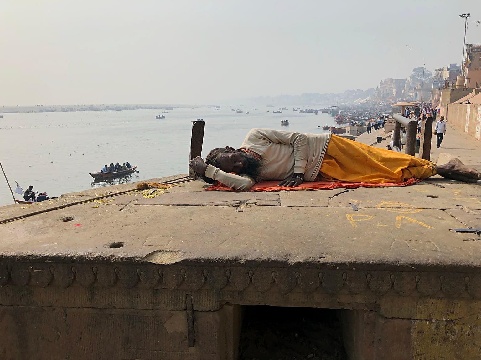 A man rests in the foreground of the ghats while the Ganges and small boats extend behind him in Varanasi, 2019