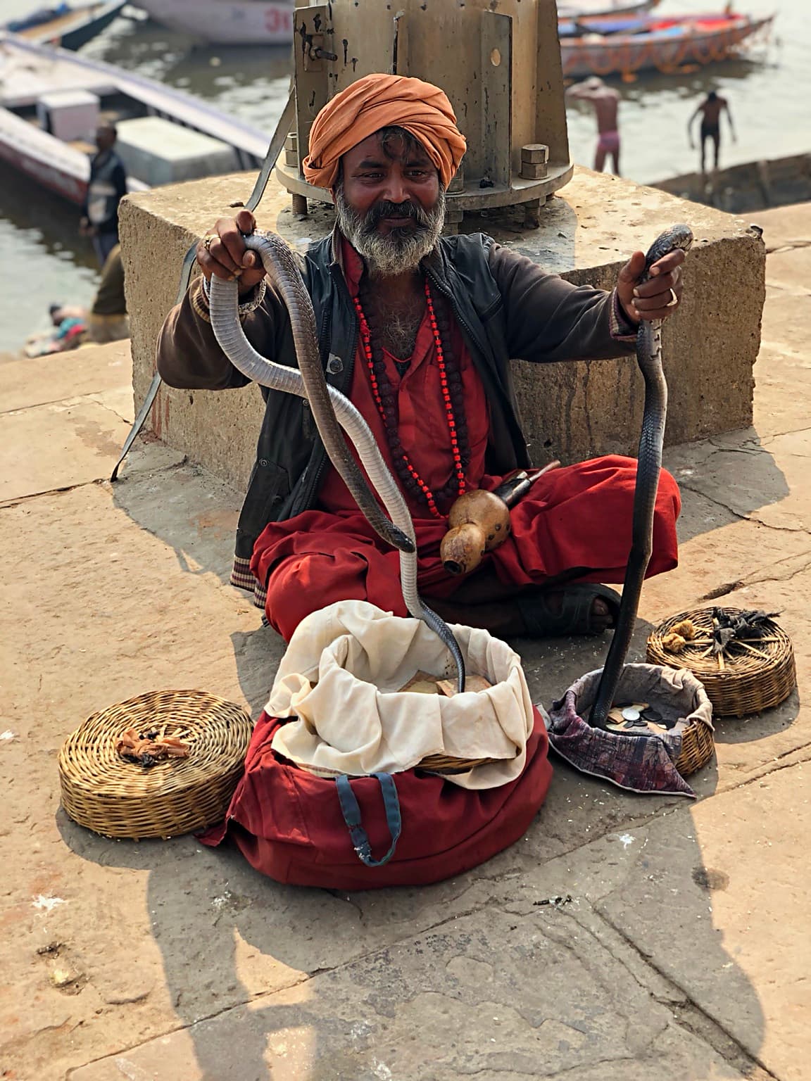 A seated snake charmer looks toward the camera beside baskets and two raised cobras on the ghats of Varanasi, 2019
