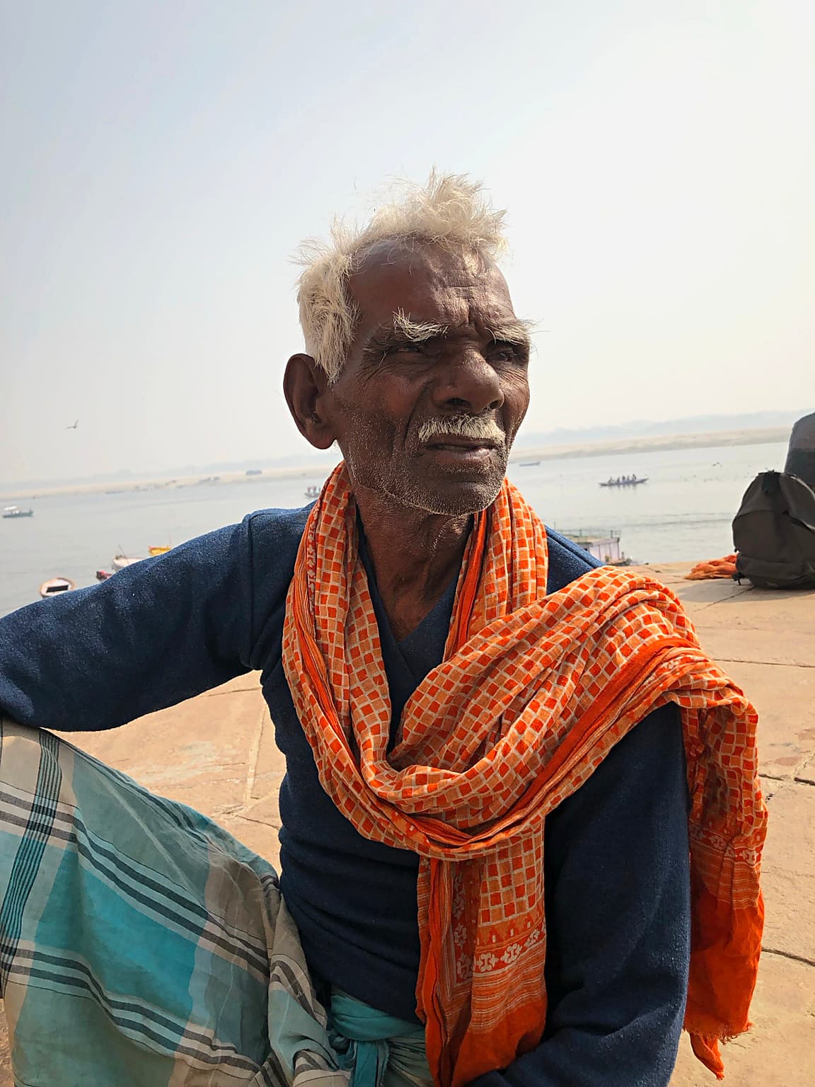 An older man with white hair, a lined face and an orange scarf looks away from the camera in Varanasi, 2019