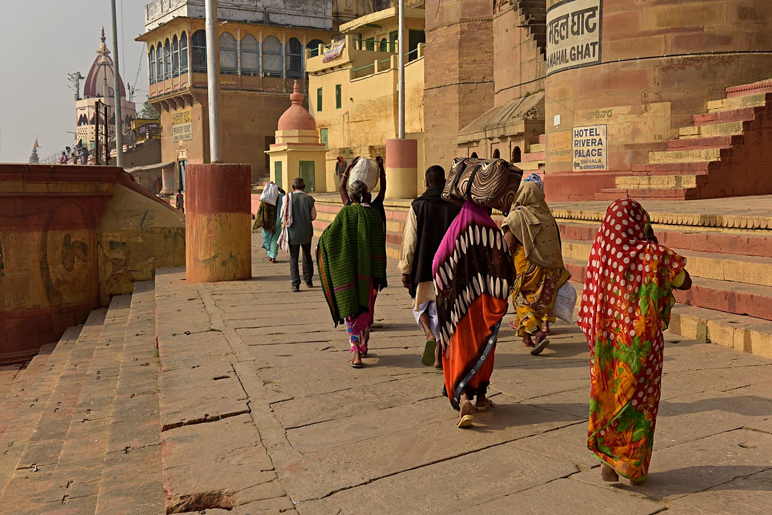 Women walk away through a ghat-side passage framed by painted architecture and signs in Varanasi, 2019