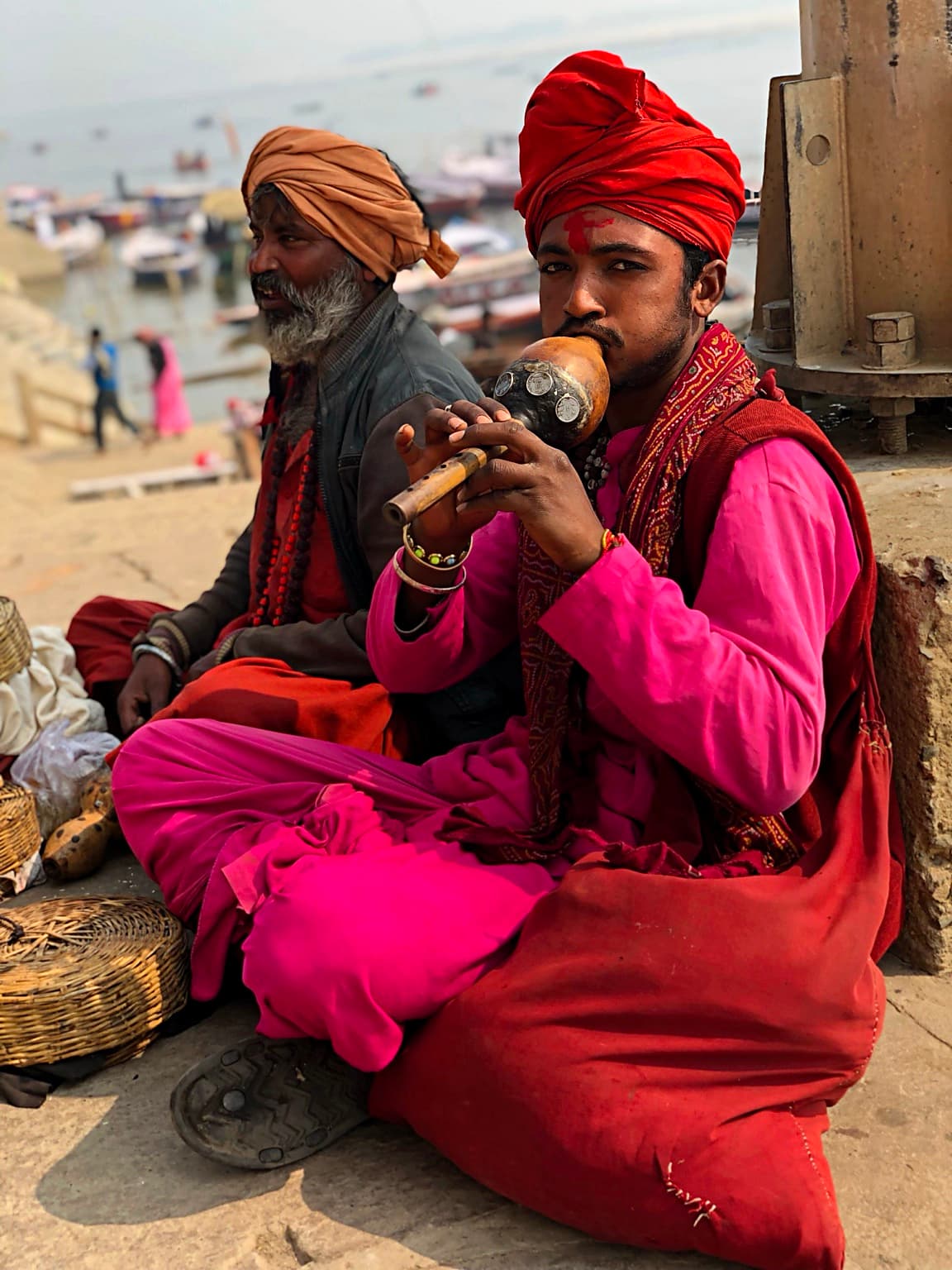 A young musician plays a pungi while an older snake charmer sits behind him on the ghats of Varanasi, 2019