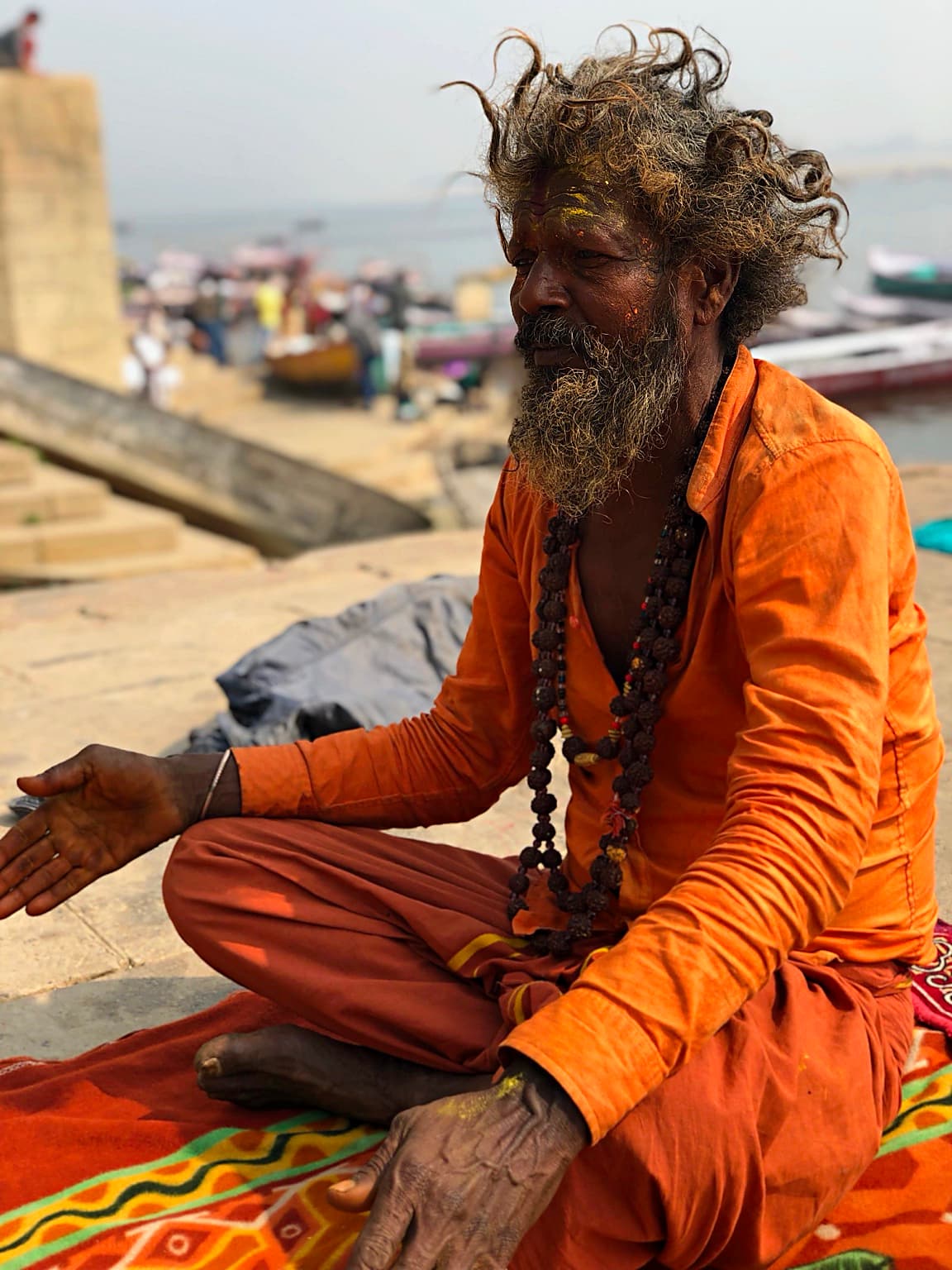 An orange-robed sadhu with long expressive hair is seen in profile on the ghats of Varanasi, 2019