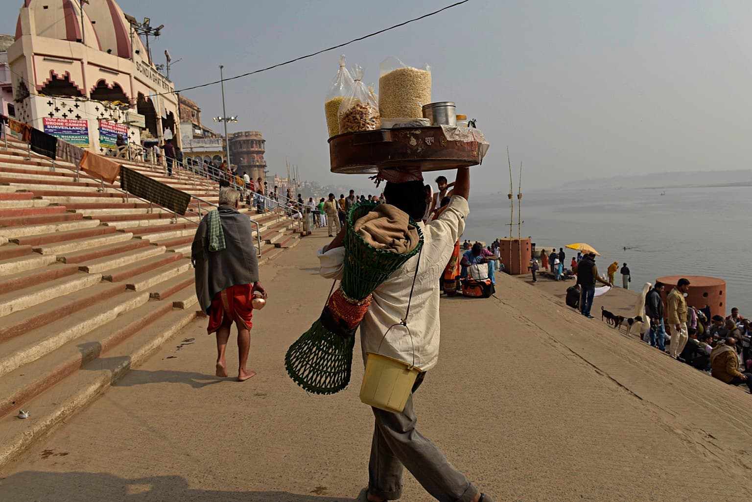 A vendor walks along the ghats carrying a large tray and baskets, with riverfront architecture behind him in Varanasi, 2019