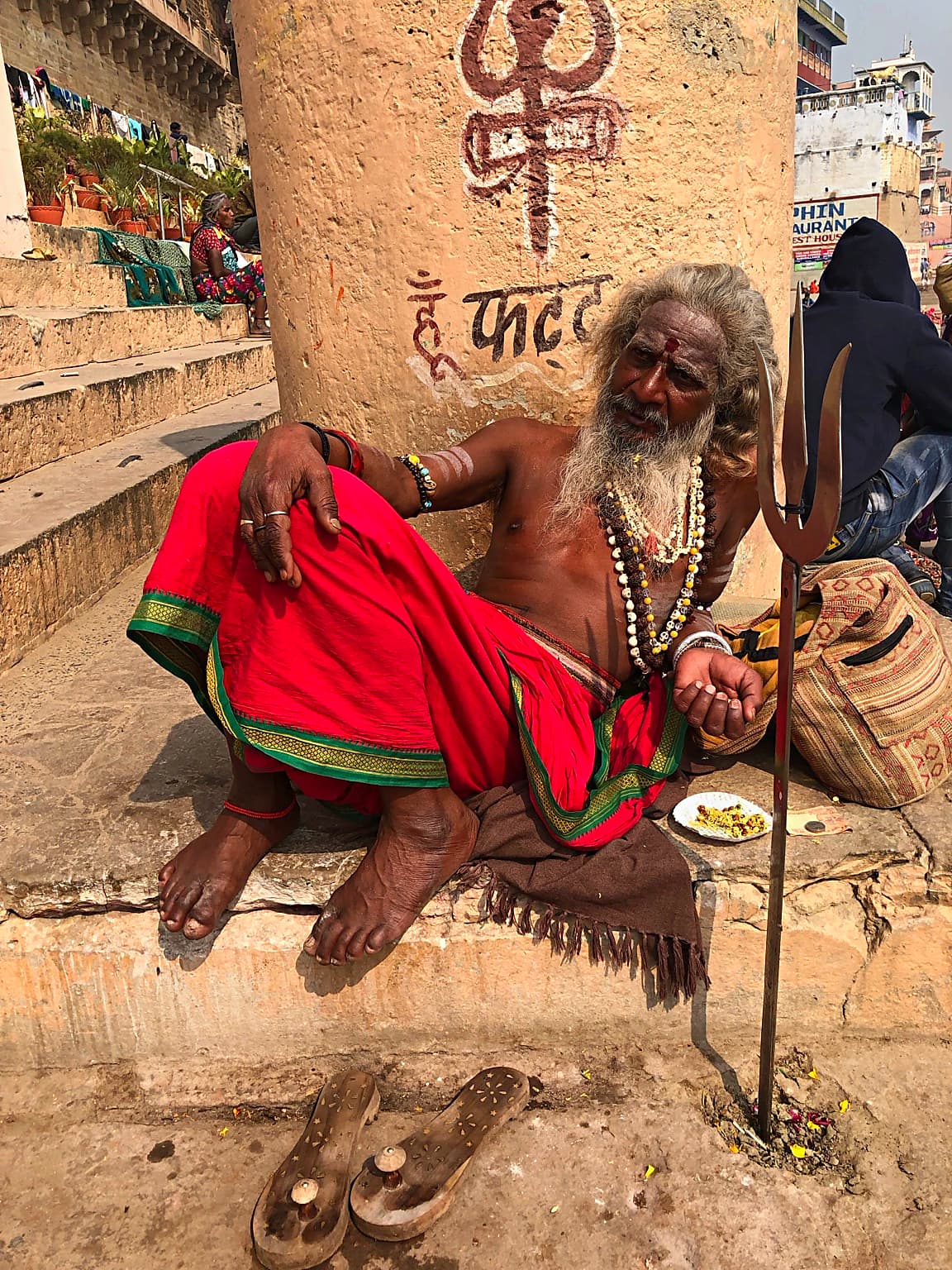 A seated sadhu with a long white beard, red cloth and trident is photographed within the ghat environment in Varanasi, 2019