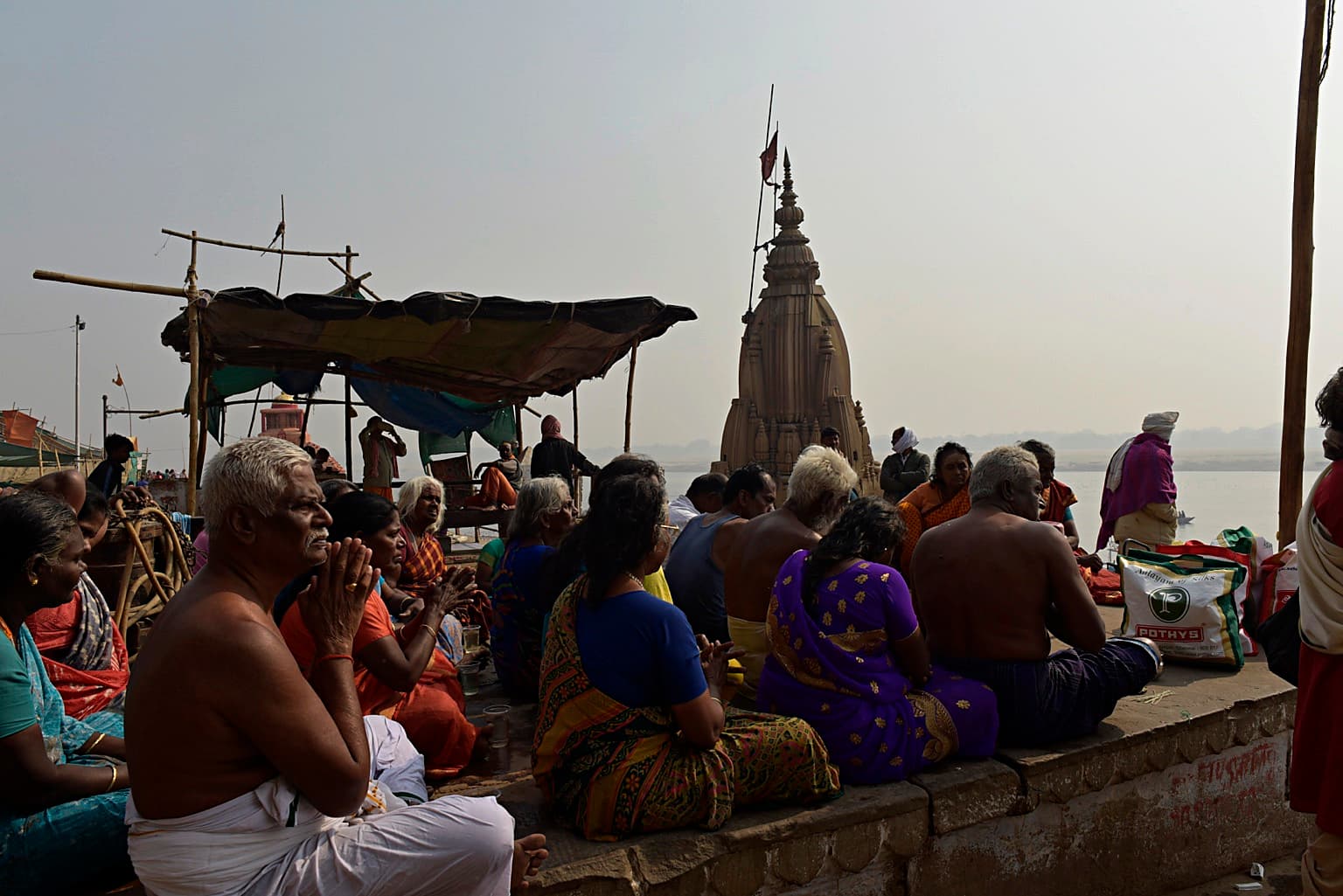 People gather around vessels and offerings on the ghats with temple architecture behind them in Varanasi, 2019