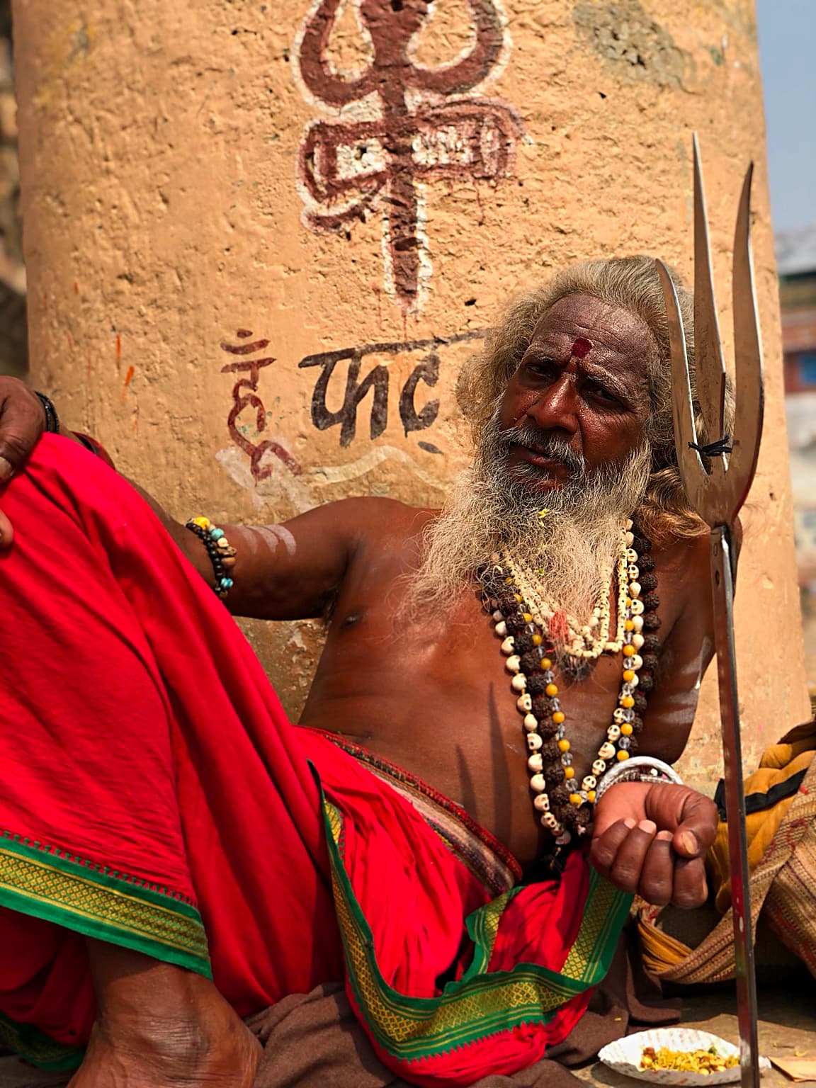 A long-bearded sadhu in red cloth sits beside a trident in a close portrait on the ghats of Varanasi, 2019