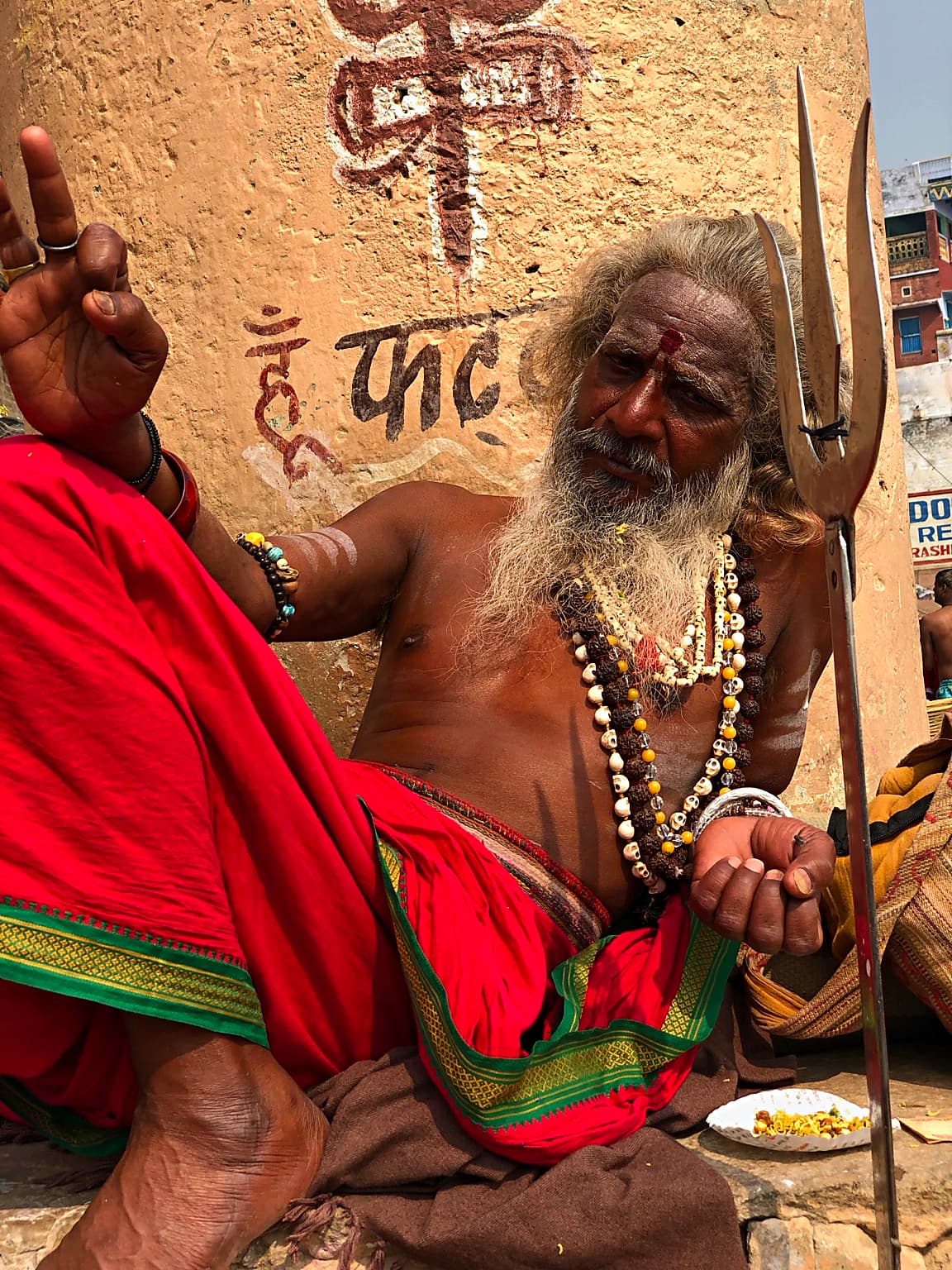 A long-bearded sadhu raises one hand while seated beside his trident on the ghats of Varanasi, 2019