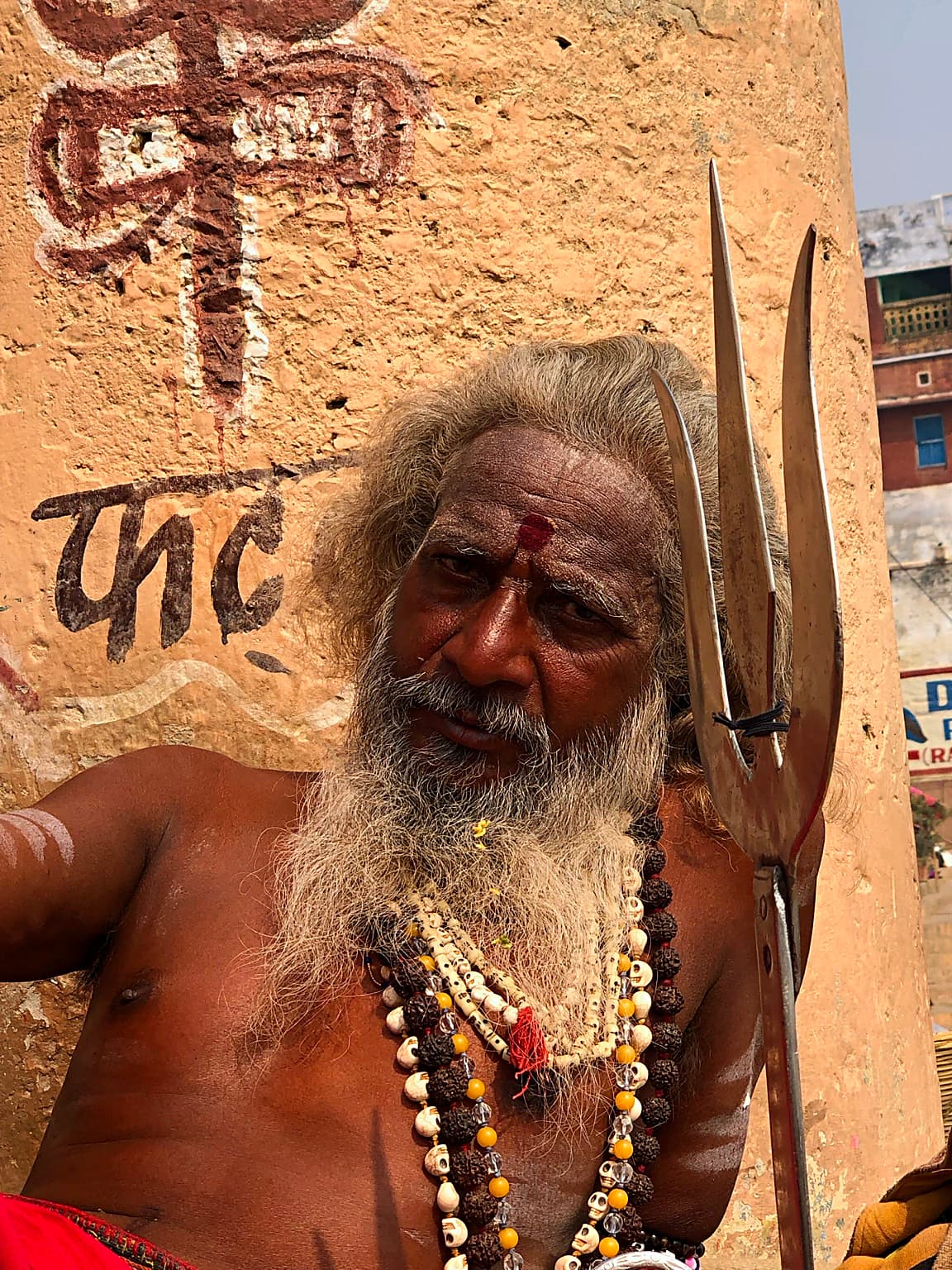 A tight portrait frames a long-bearded sadhu with layered beads and a trident in Varanasi, 2019