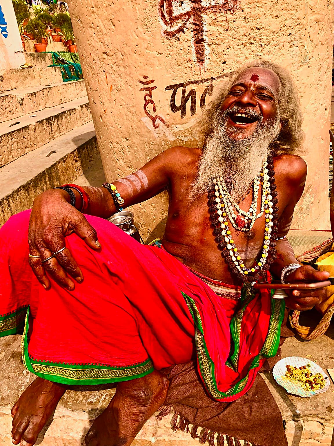 A long-haired sadhu laughs openly while seated in bright red cloth on the ghats of Varanasi, 2019