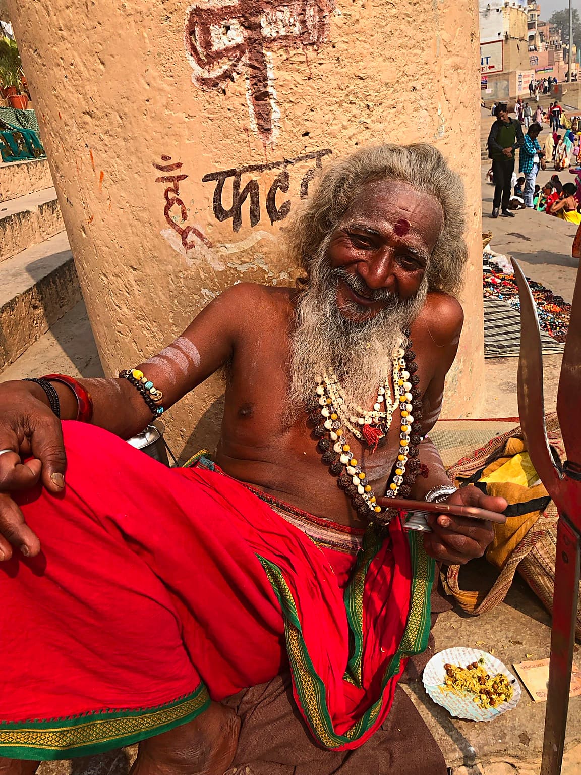 A long-haired sadhu smiles while seated in red cloth beside his belongings on the ghats of Varanasi, 2019