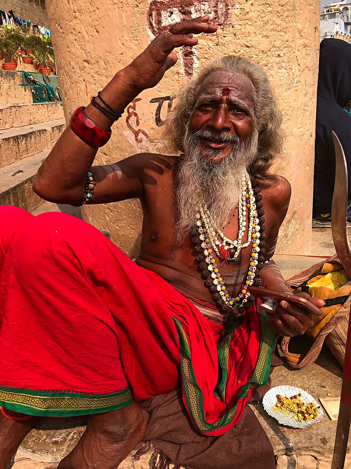 A smiling sadhu raises one arm while seated in vivid red cloth on the ghats of Varanasi, 2019